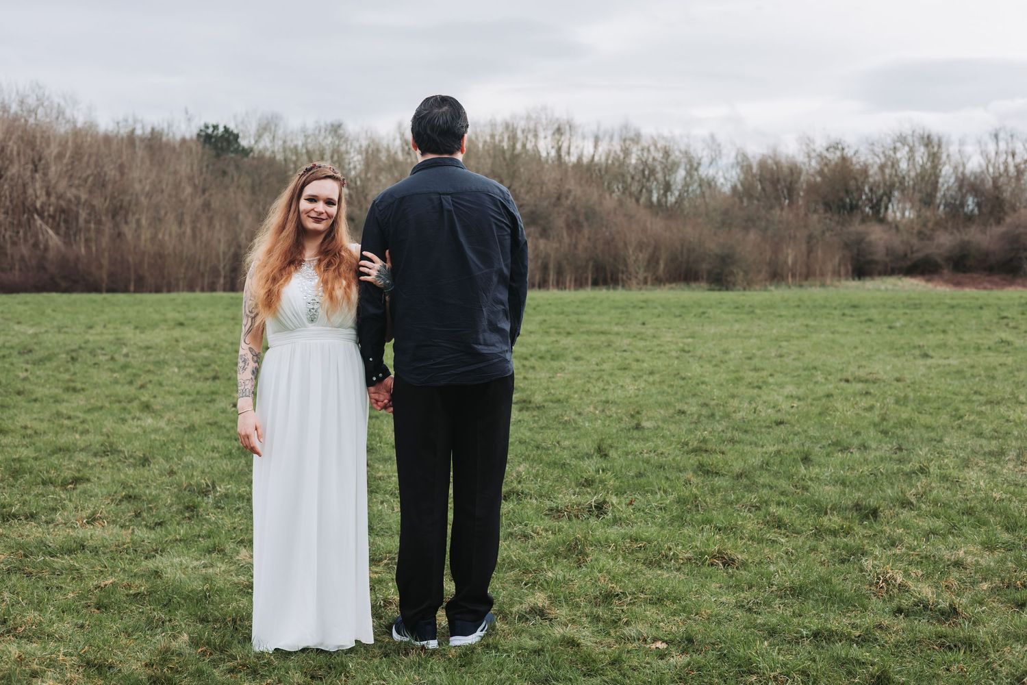 A couple stands together in a green field wearing formal attire on their wedding day.