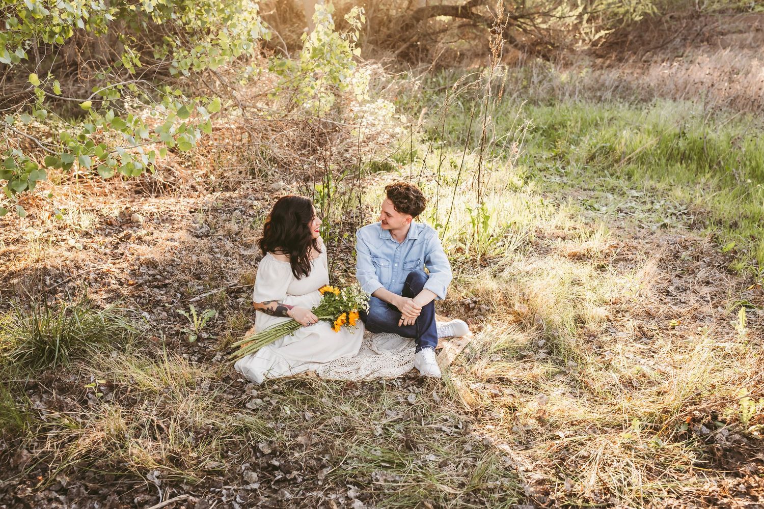 A couple sits together on a blanket in a sun-dappled forest clearing during an outdoor photoshoot session.