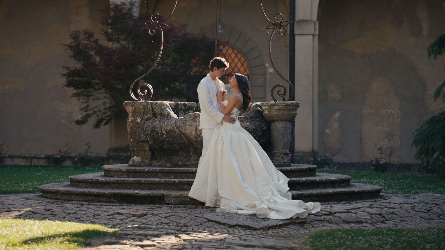 A bride and groom share a romantic embrace near a stone fountain in an elegant outdoor courtyard setting.