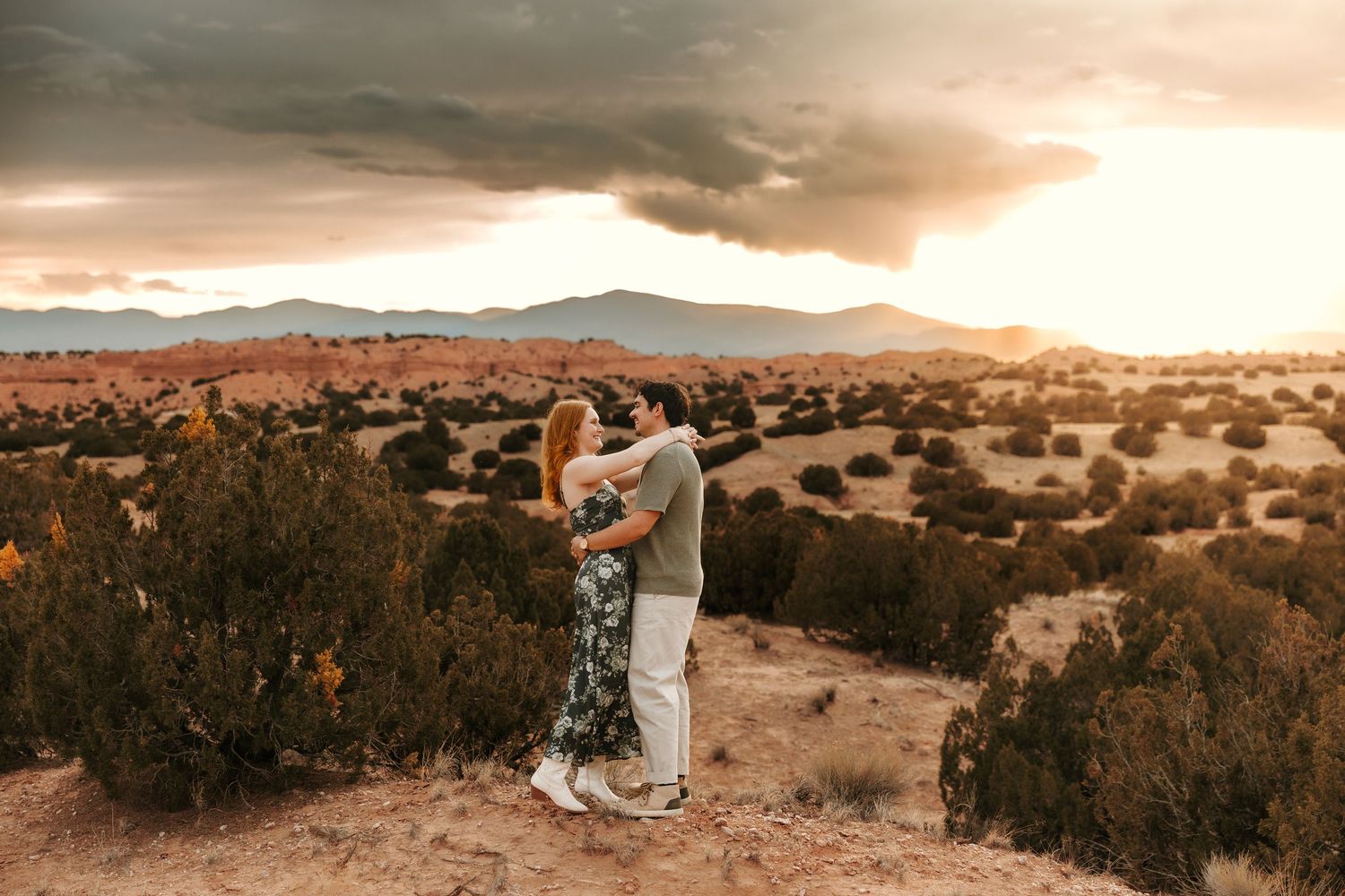 Couple embracing at sunset in a desert landscape with dramatic storm clouds and red rock formations in the background.