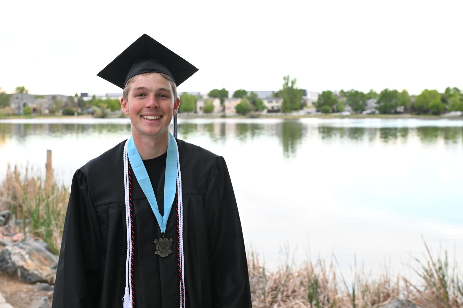Smiling graduate in black cap and gown with blue medal stands by a lake on a cloudy day.