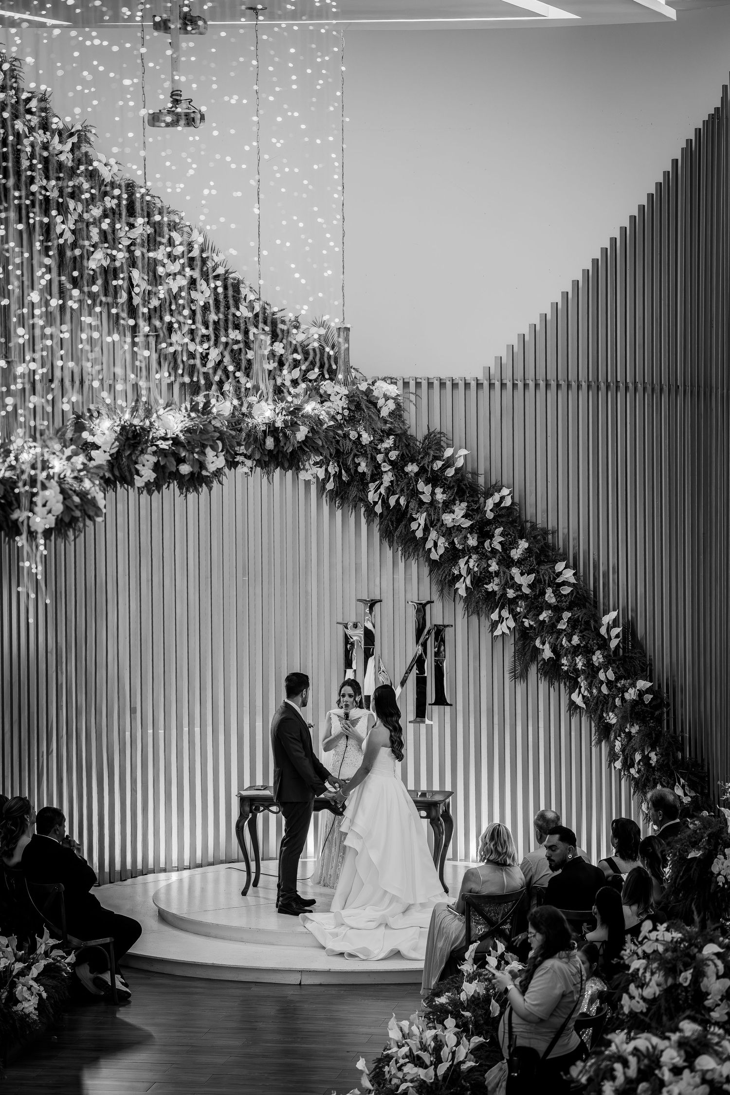 Black and white wedding ceremony photo with couple exchanging vows under floral arch with fairy lights.