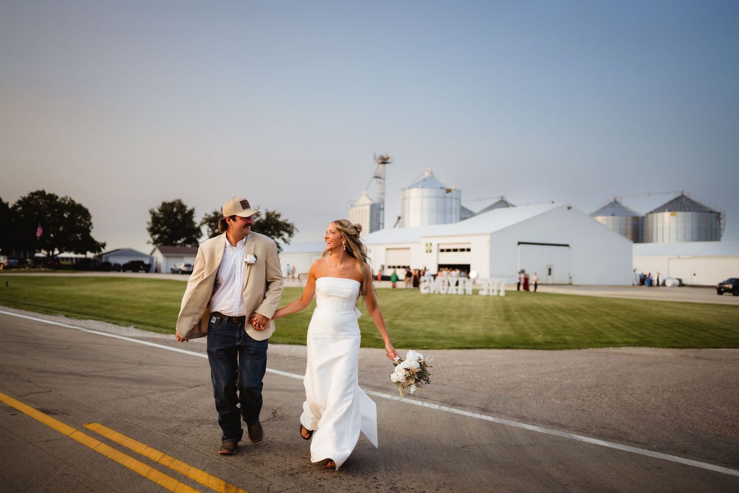 Bride and groom walking hand-in-hand down a rural road past white grain silos on a sunny wedding day.
