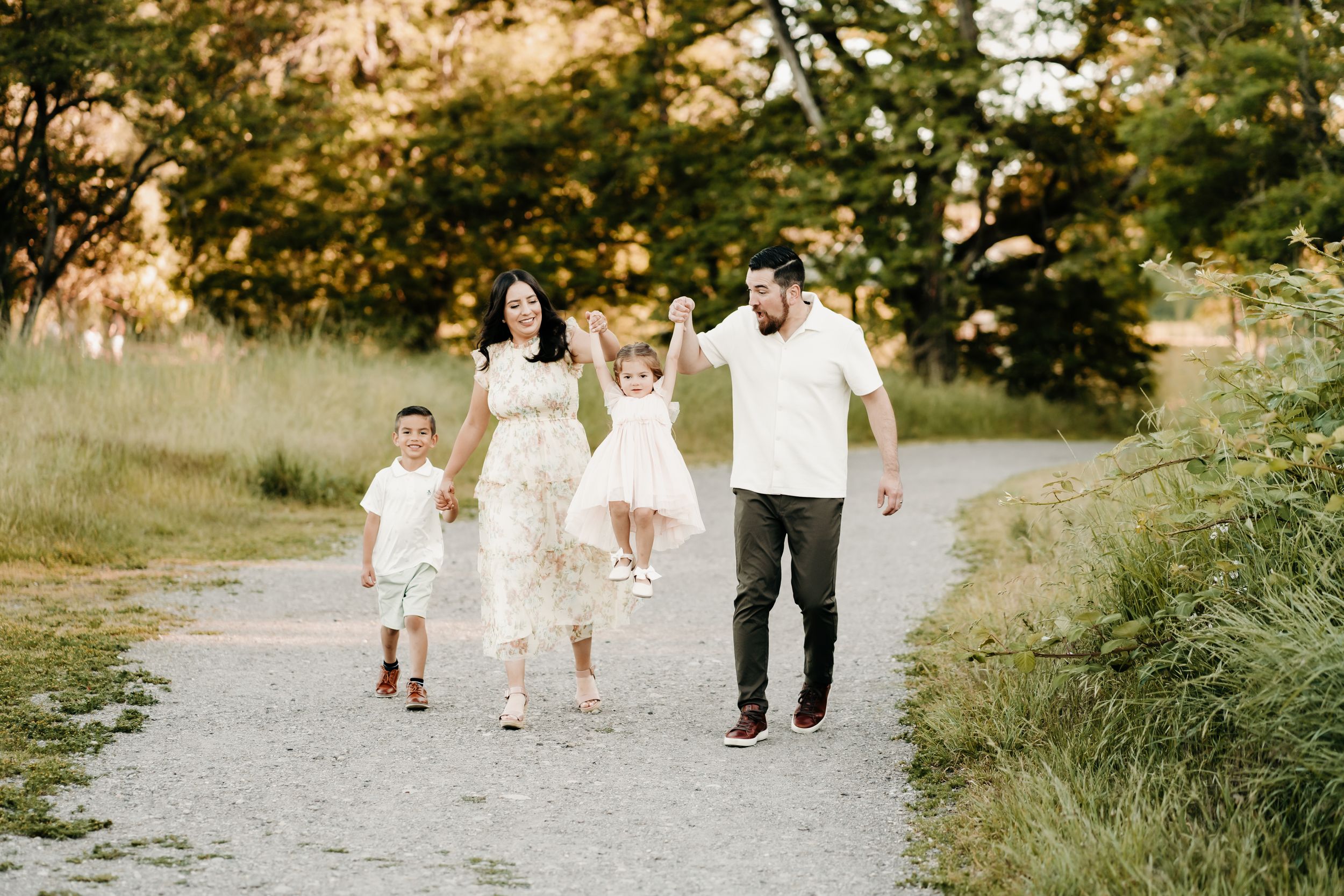 Family of four walking on a gravel path in a sunny park, parents swinging young children between them during family photos in Puyallup, Wa.