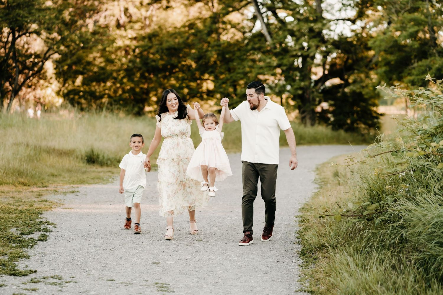Family of four walking on a gravel path in a sunny park, parents swinging young children between them during family photos in Puyallup, Wa.