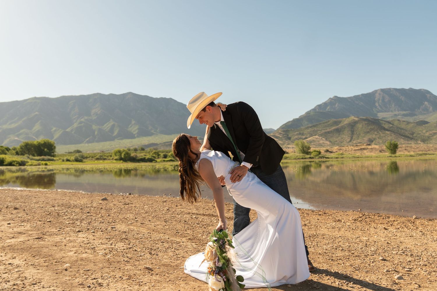 Couple sharing a romantic dip kiss at a mountain lake during outdoor wedding ceremony in cowboy hat.