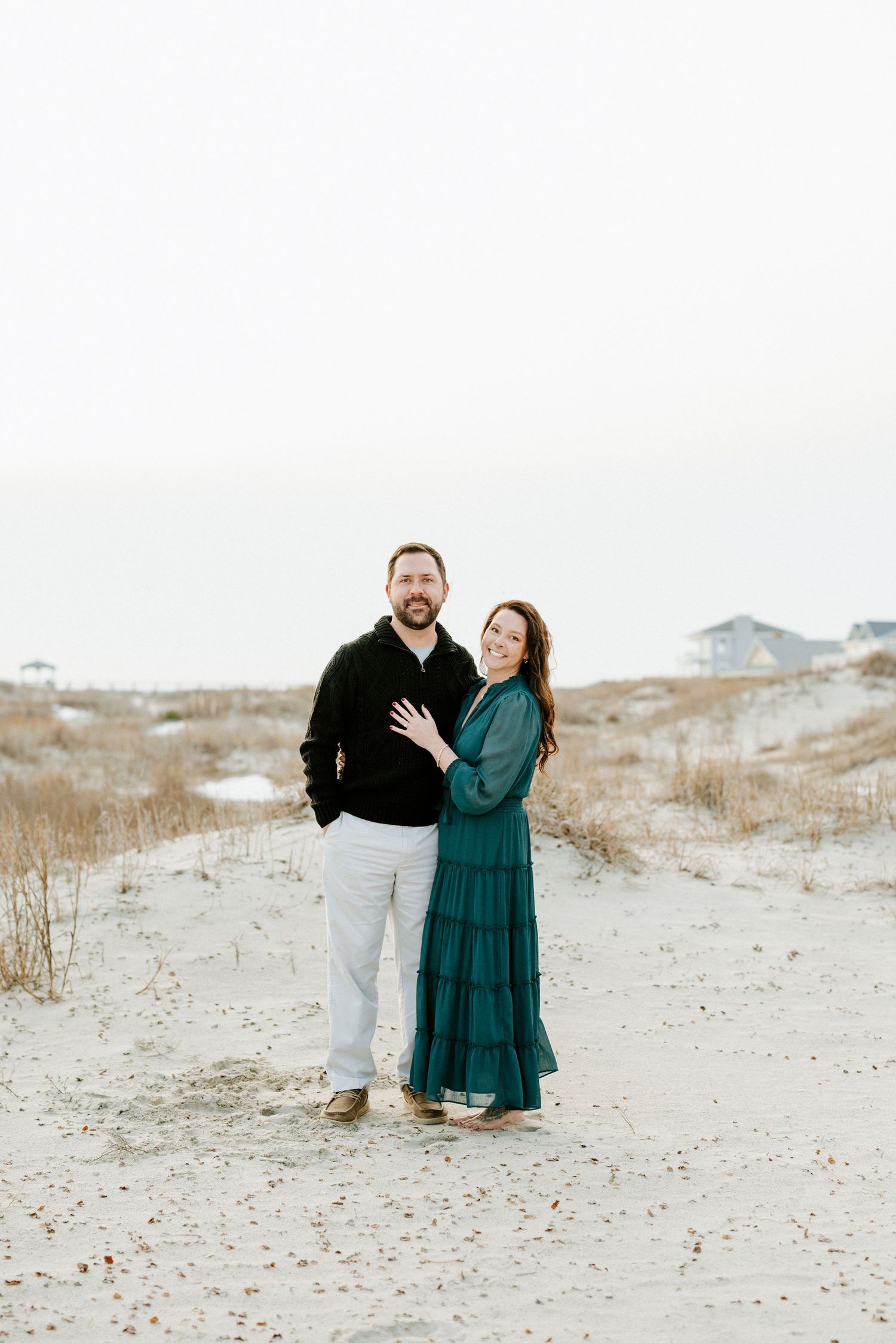 A couple stands together on sandy dunes at sunset wearing dark clothing and embracing in a romantic moment.