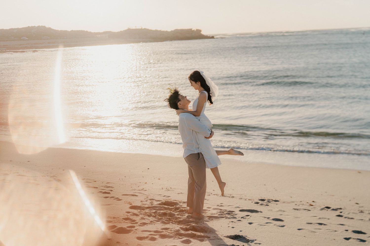 A couple embraces on a sandy beach at sunset with ocean waves and golden sunlight in the background.