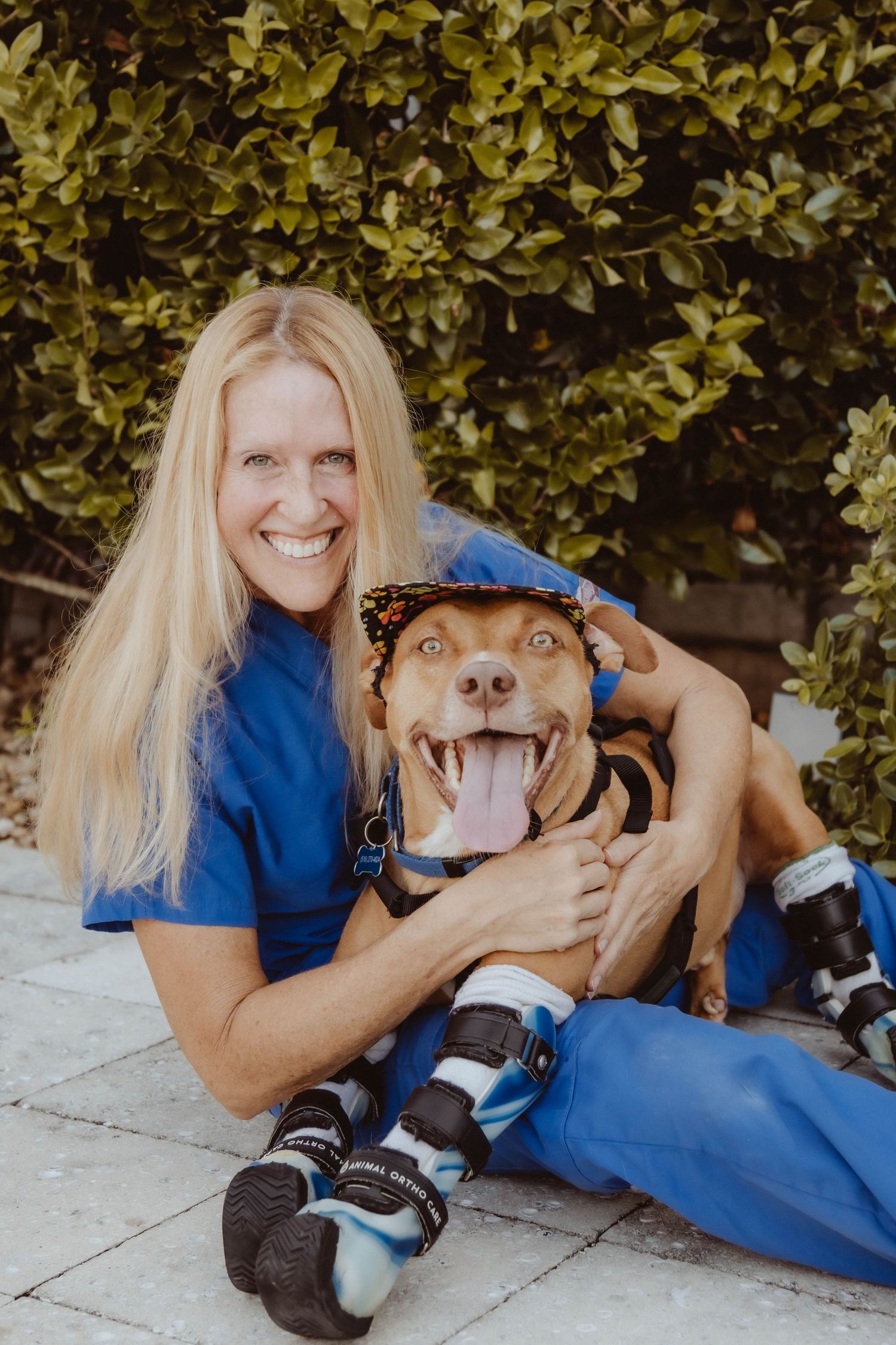 Smiling blonde woman in blue scrubs hugging a happy brown dog wearing a hat and boots outdoors.