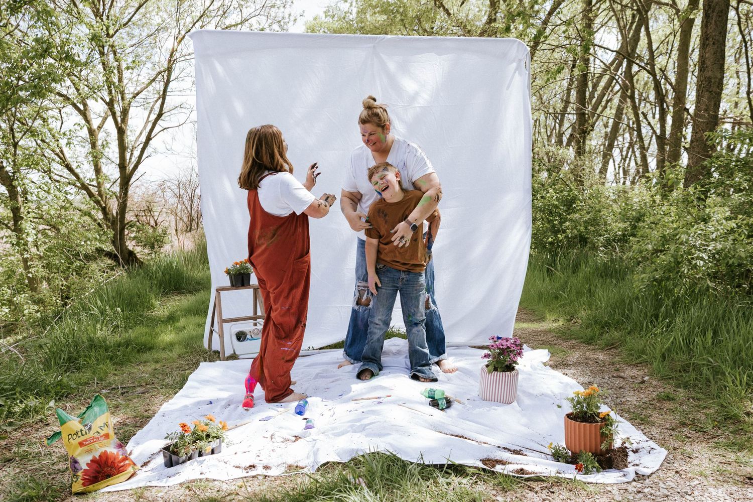 Two women and a child pose in front of a white backdrop outdoors, surrounded by plants and props in a natural setting.