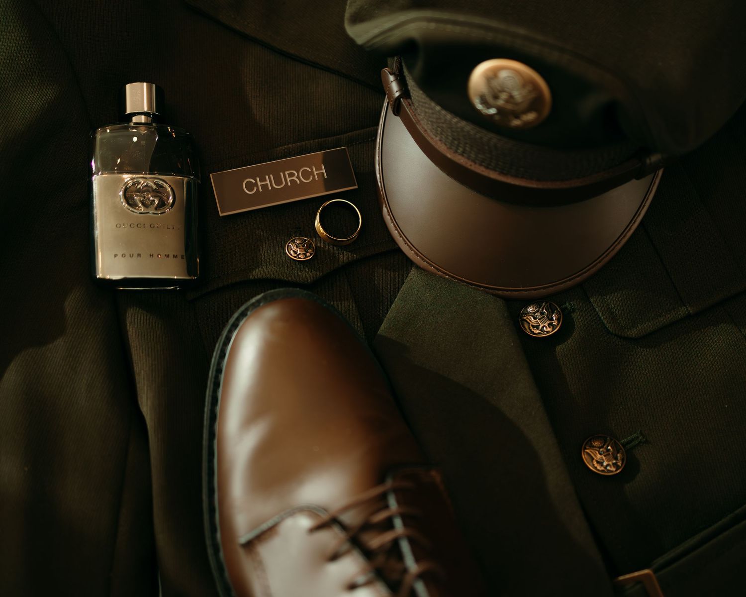 Military uniform flat lay with Gucci cologne, brown Oxford shoes, name tag, and officer cap on dark fabric.