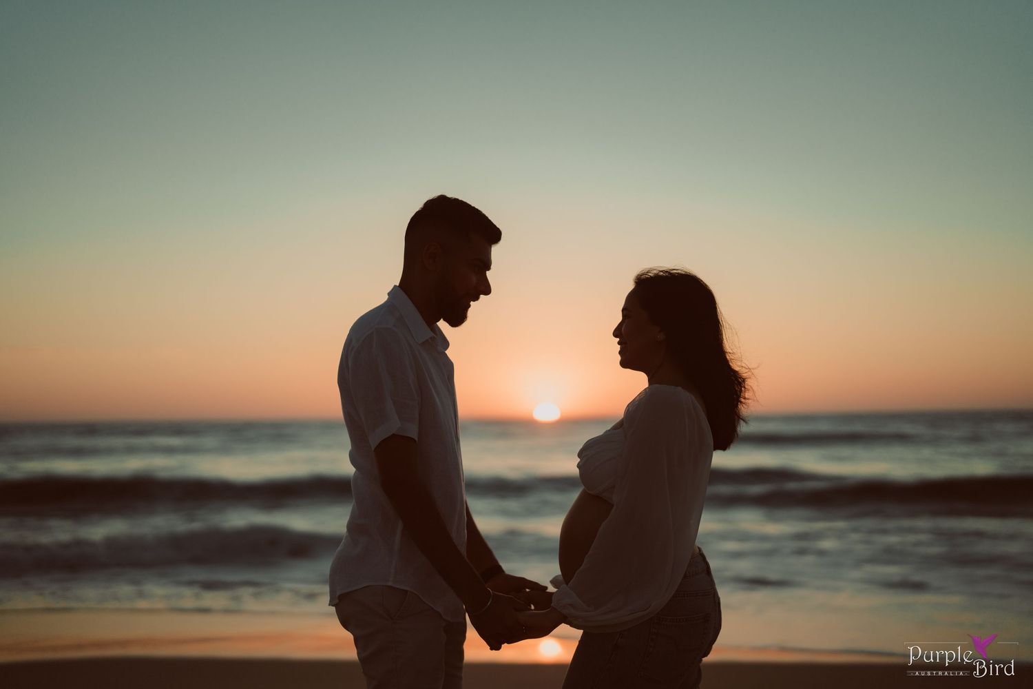 Silhouette of an expecting couple holding hands on a beach at sunset, with waves and a glowing orange sky behind them.
