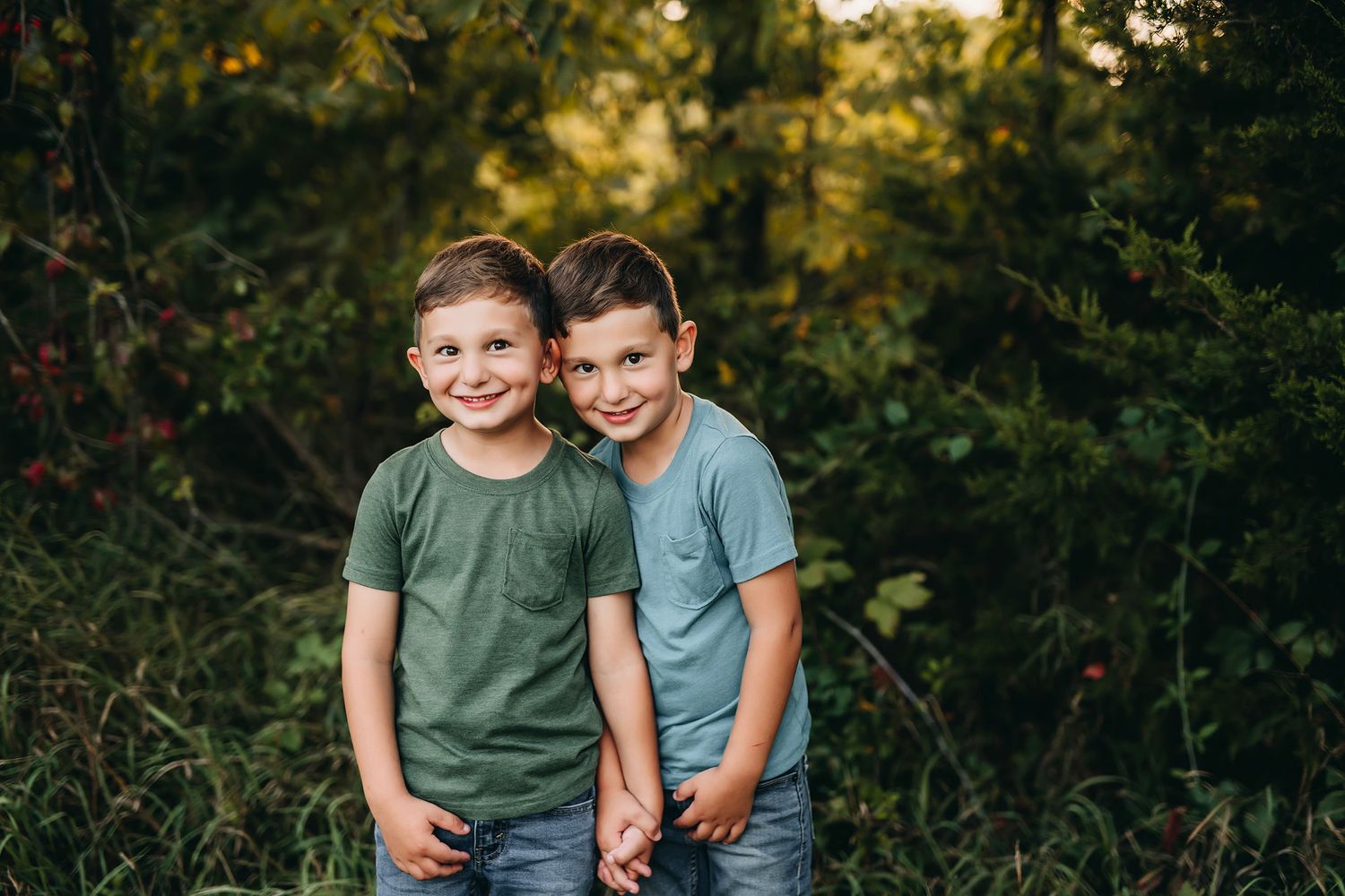 Two smiling boys in matching green shirts pose together outdoors surrounded by lush green foliage.