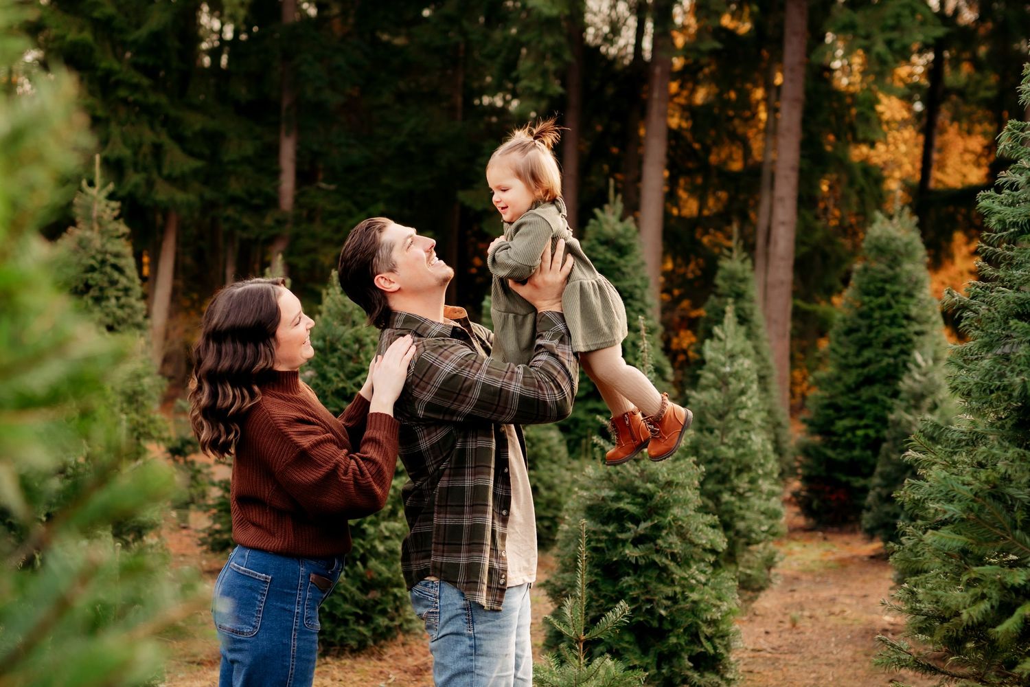 Family choosing a Christmas tree at an outdoor tree farm, father lifting toddler while mother watches joyfully.