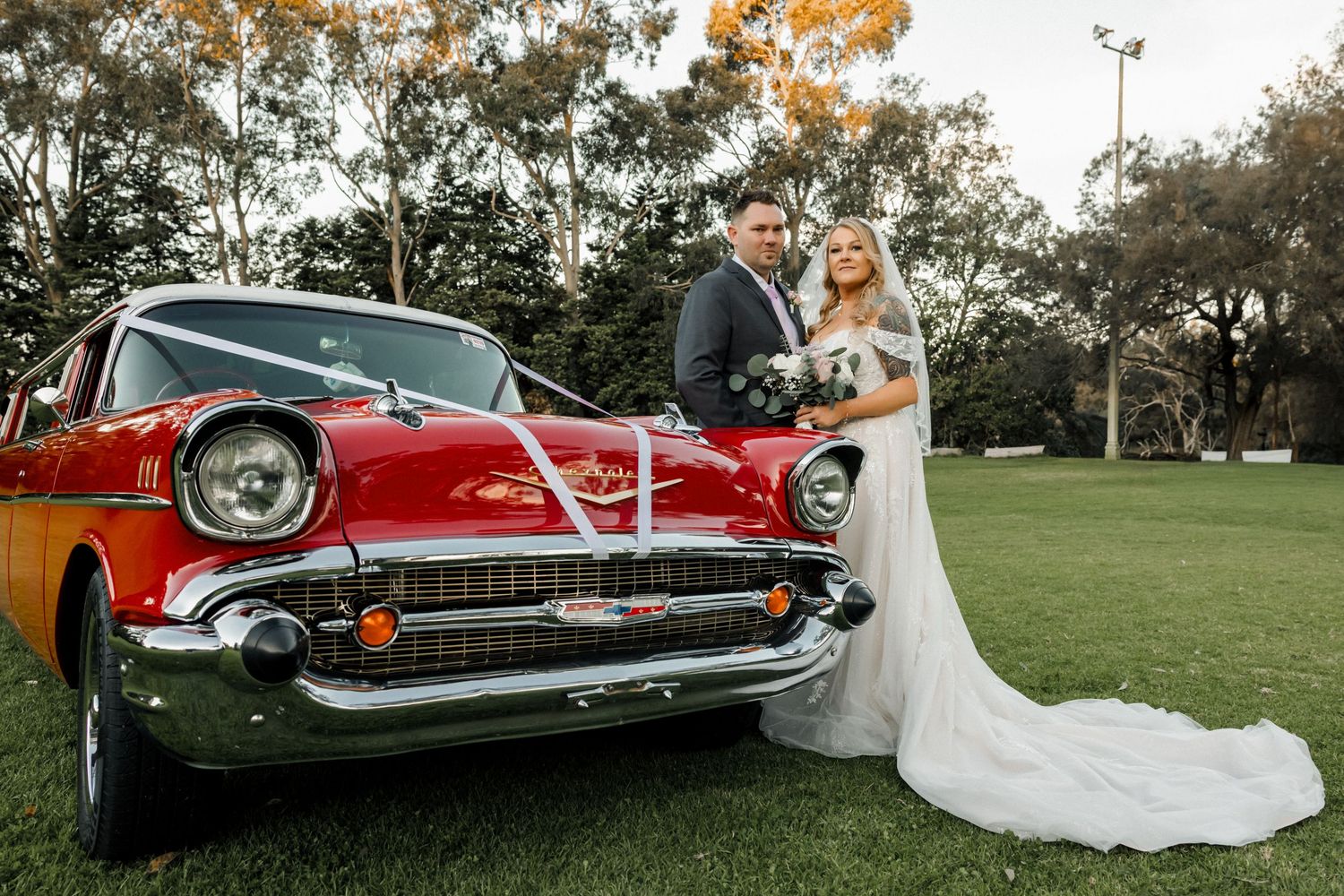 Bride and groom standing beside a classic red 1957 Chevrolet wedding car decorated with white ribbons.