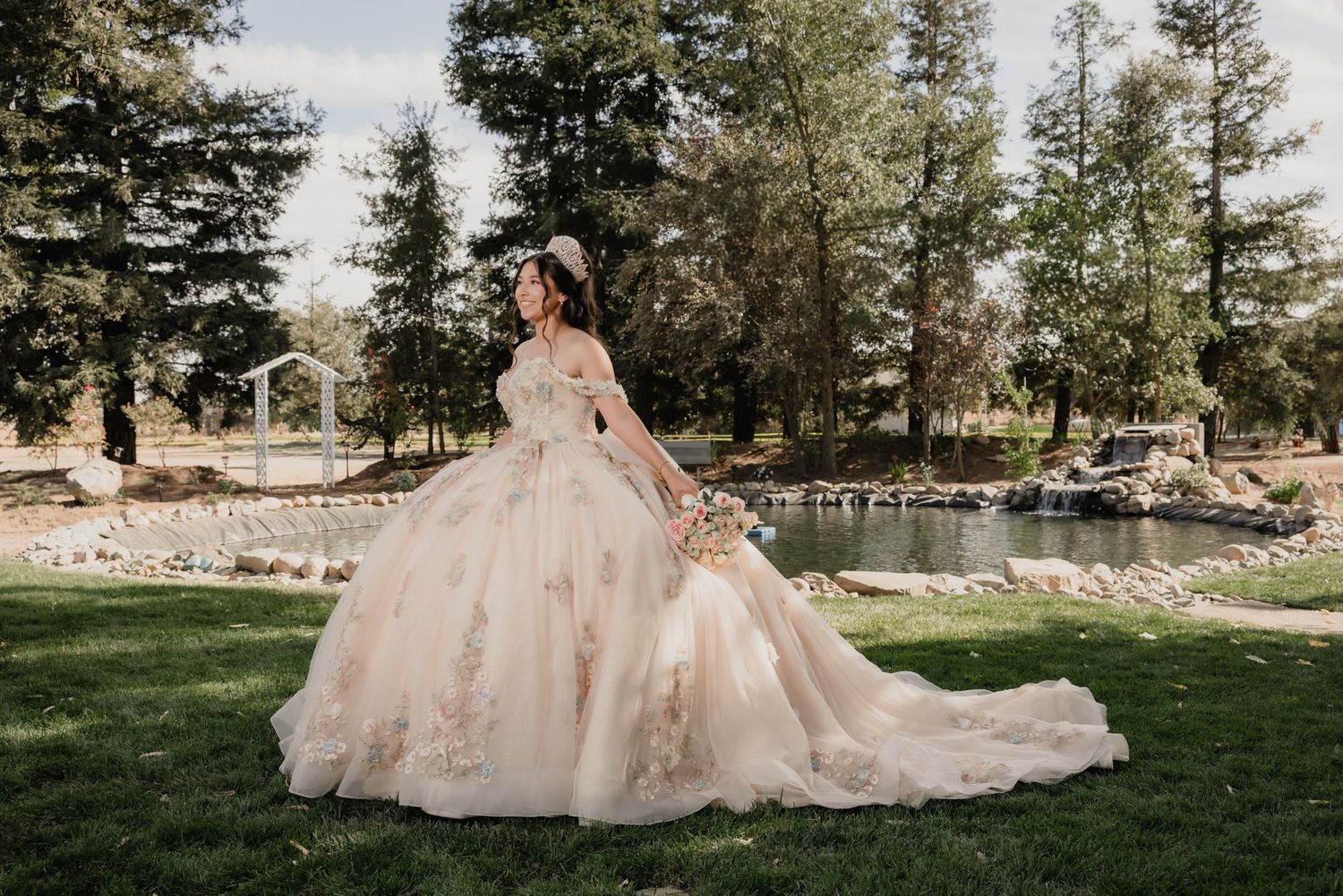 Young woman in a red ball gown and crown poses among roses for a quinceañera portrait.