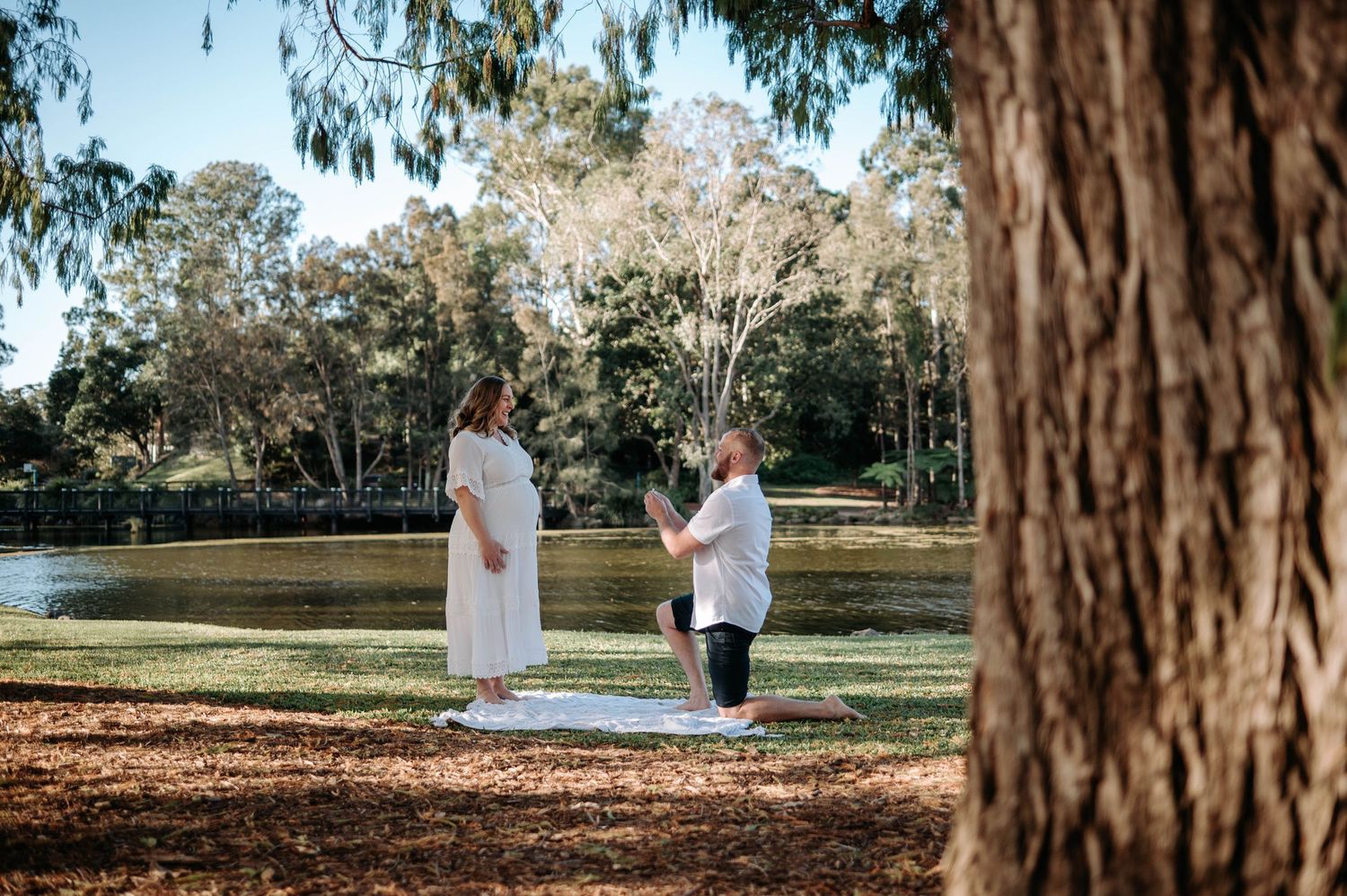 Man proposing on one knee to woman in white dress by a tranquil lake in a lush green park.