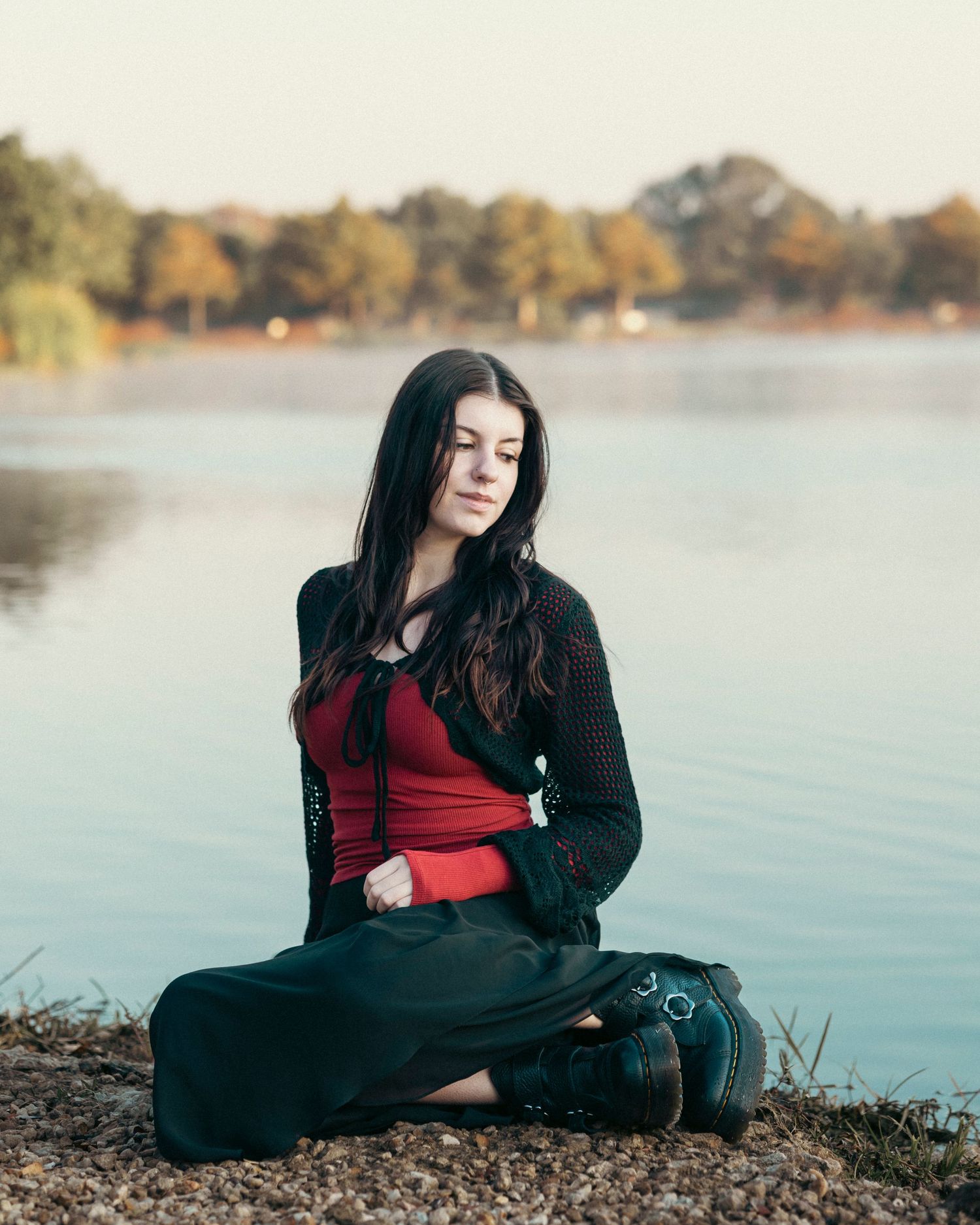 Young woman with dark hair sits peacefully by a serene lake, wearing a red top and black cardigan.