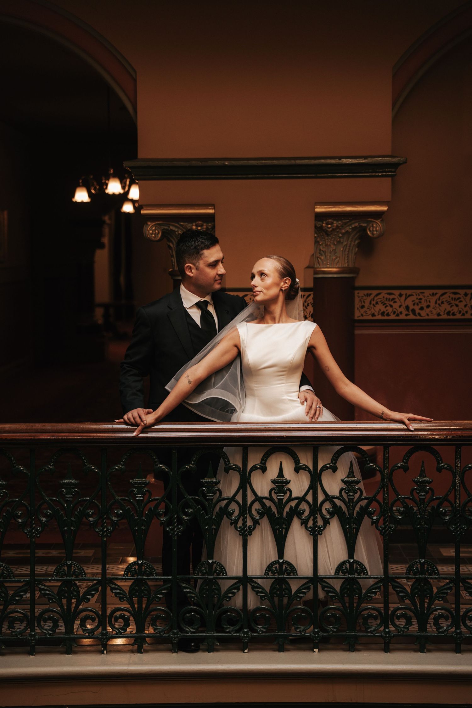 Elegant wedding couple in formal attire sharing a romantic moment on an ornate wrought iron balcony in a grand historic venue.