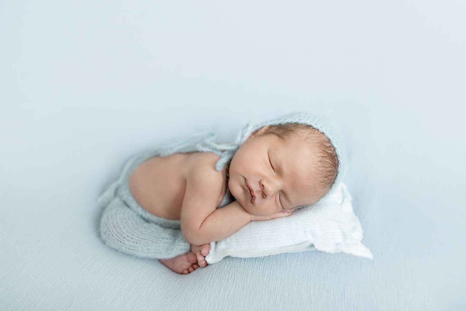 Peaceful newborn baby sleeping on a tiny white pillow, wrapped in a soft blue knit blanket on a light blue background.
