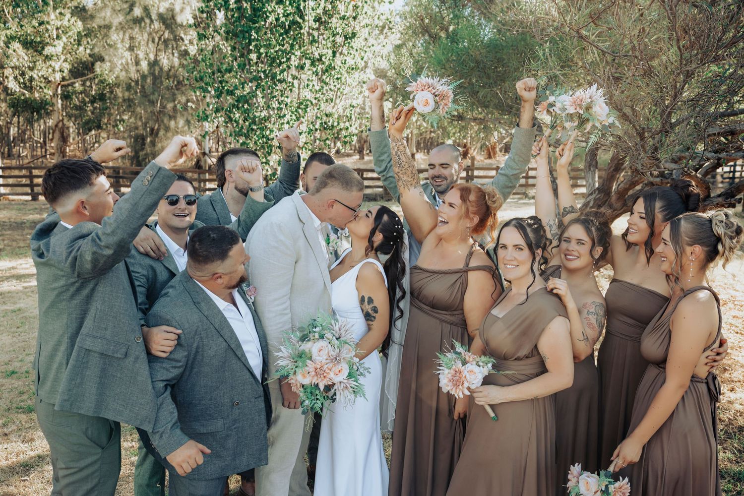 Wedding party celebrating with bride and groom kissing, bridesmaids in brown dresses holding bouquets, groomsmen cheering outdoors.