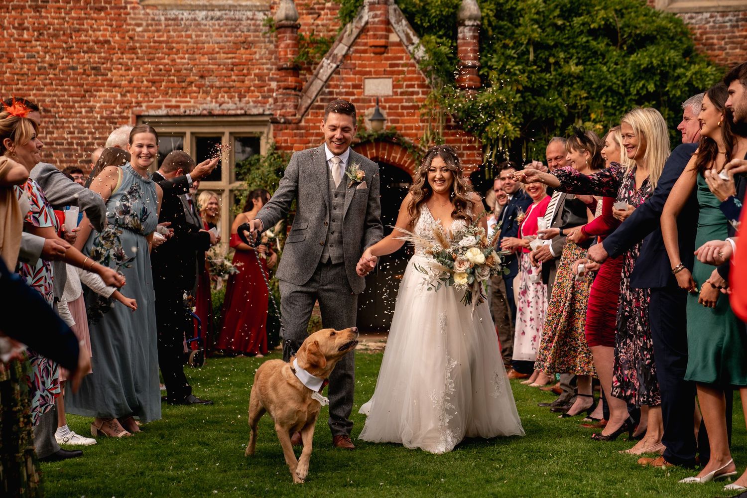 Bride and groom walk through confetti line with dog at outdoor wedding ceremony near brick venue.