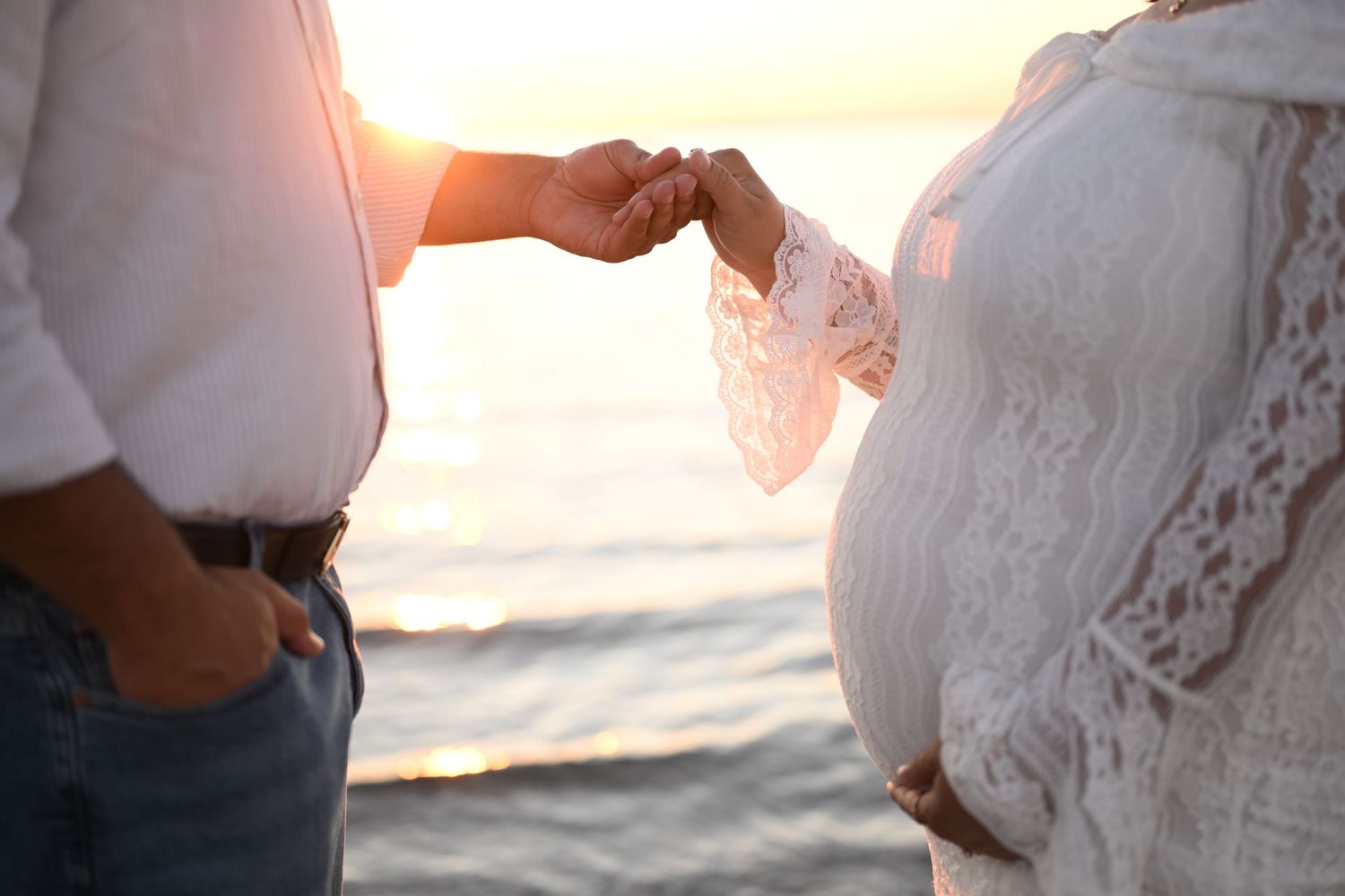 Pregnant woman in white lace dress holds hands with partner on beach at sunset.