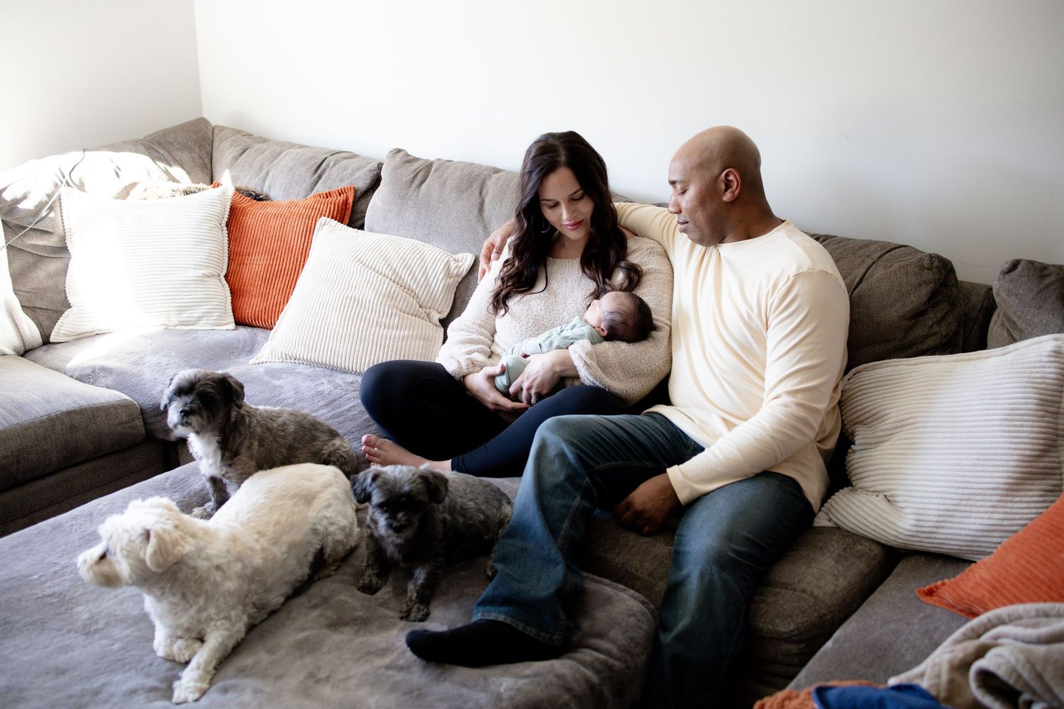 A couple sits on a gray sofa with orange pillows, holding their newborn baby while three small dogs rest on the cushions in front of them.