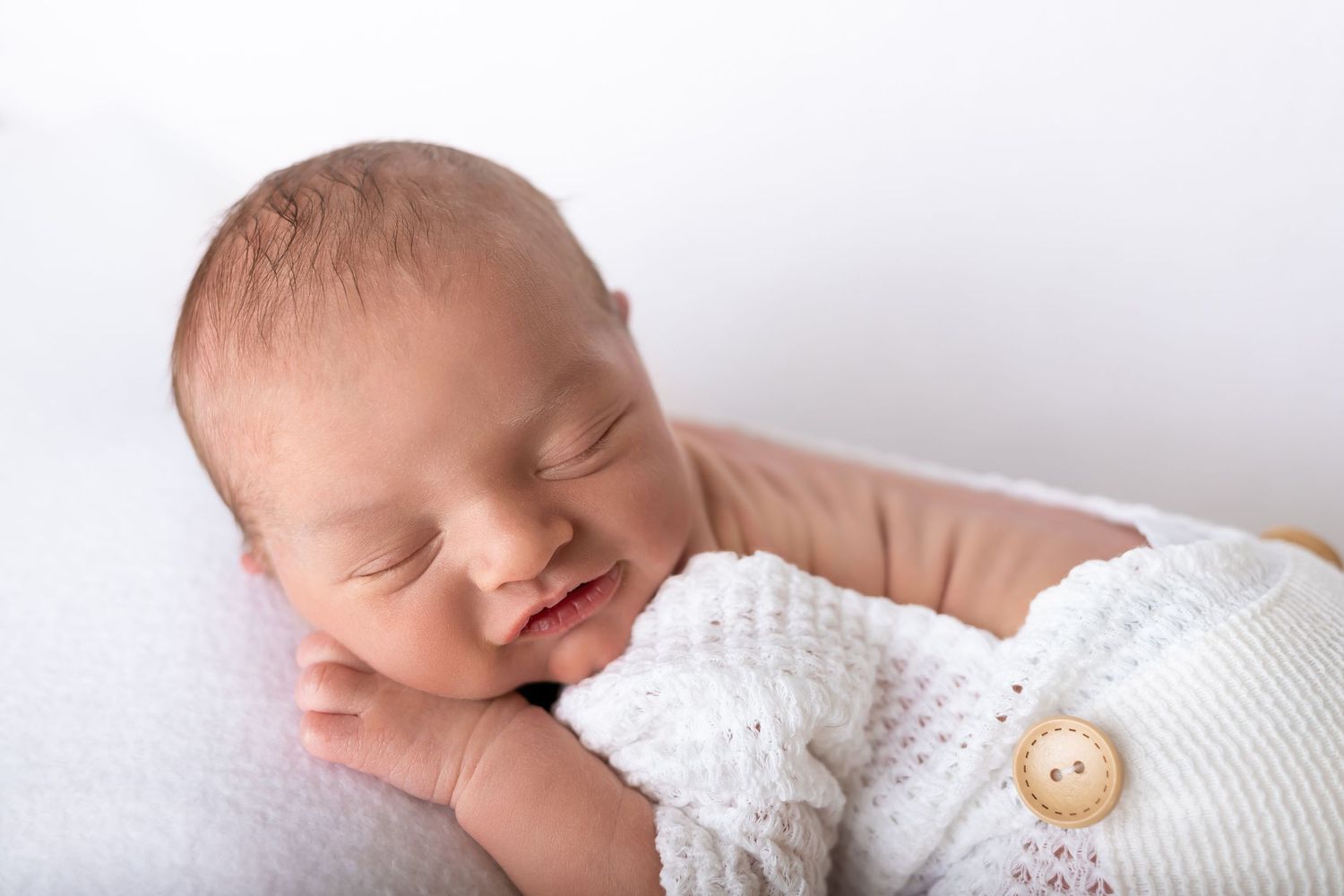 Sleeping newborn in white knit outfit smiles peacefully against light background.