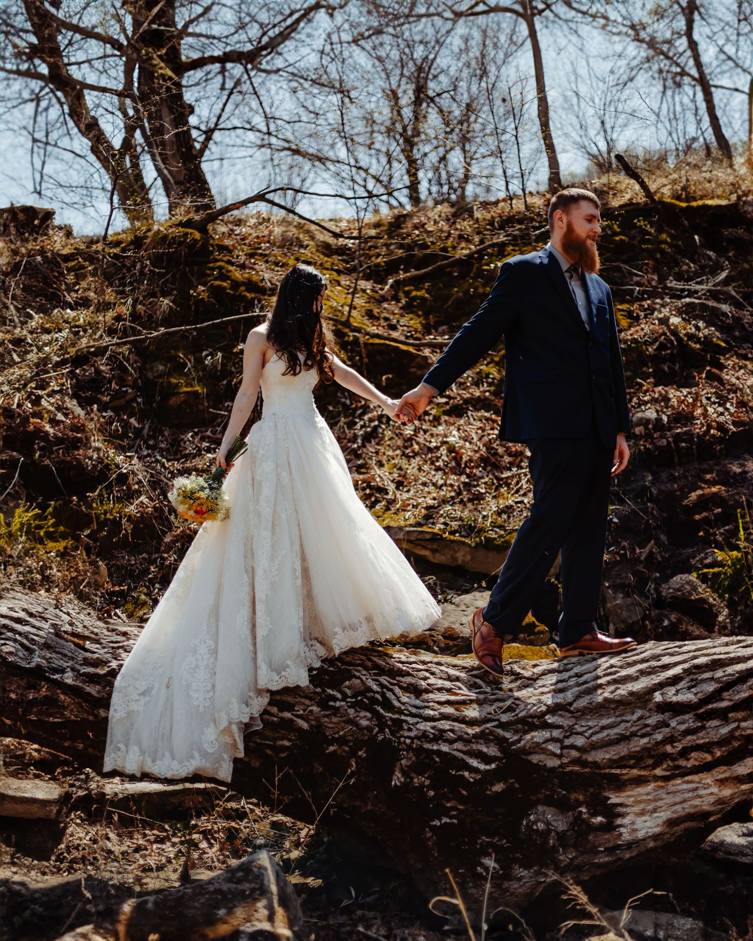 Couple in wedding attire walking across fallen tree trunk in autumn forest.