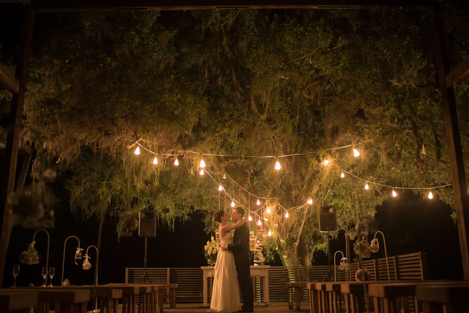 Couple shares first dance under string lights with trees in background.