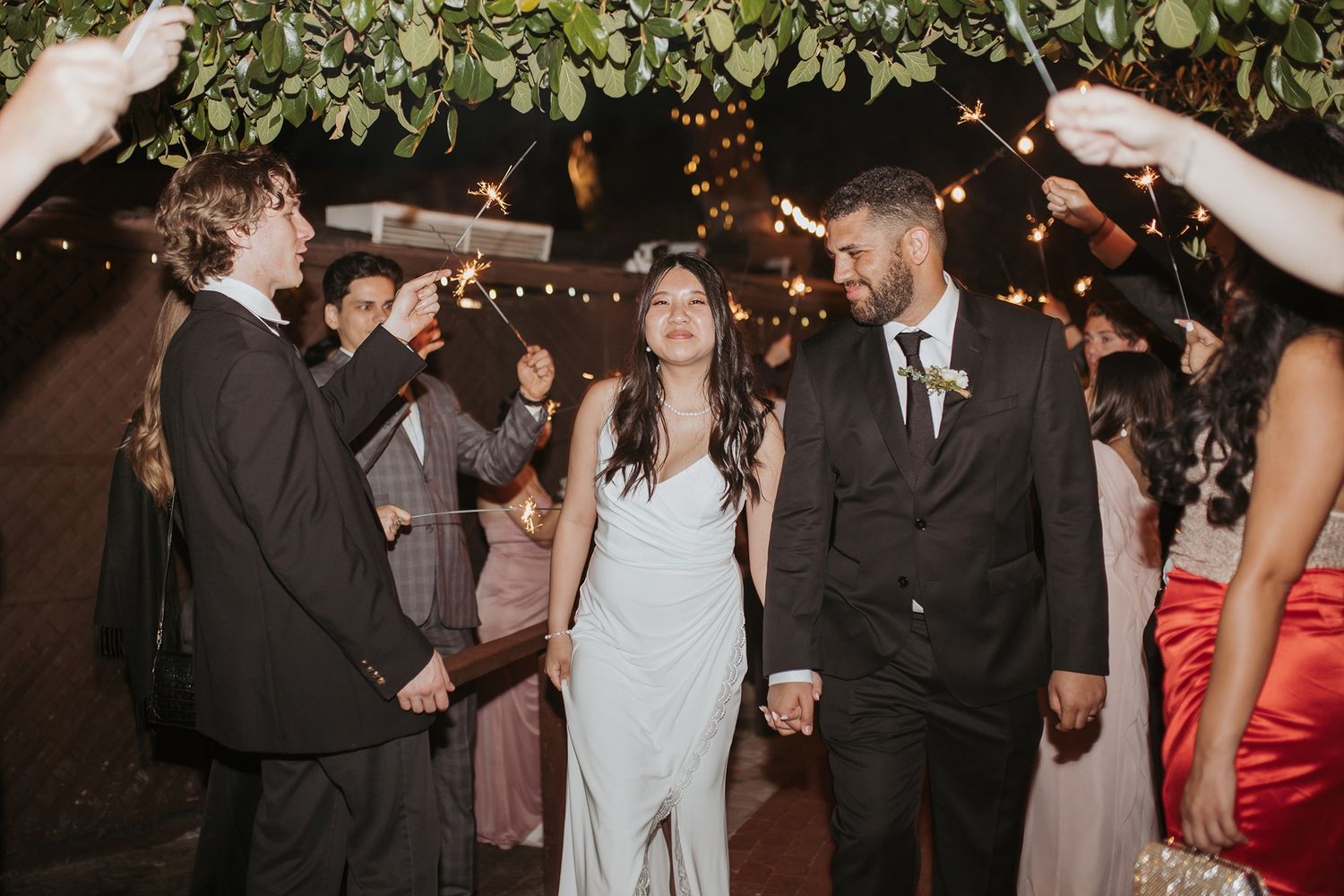 Wedding guests cheering and celebrating together at an evening reception under string lights and decorative greenery with sparklers.