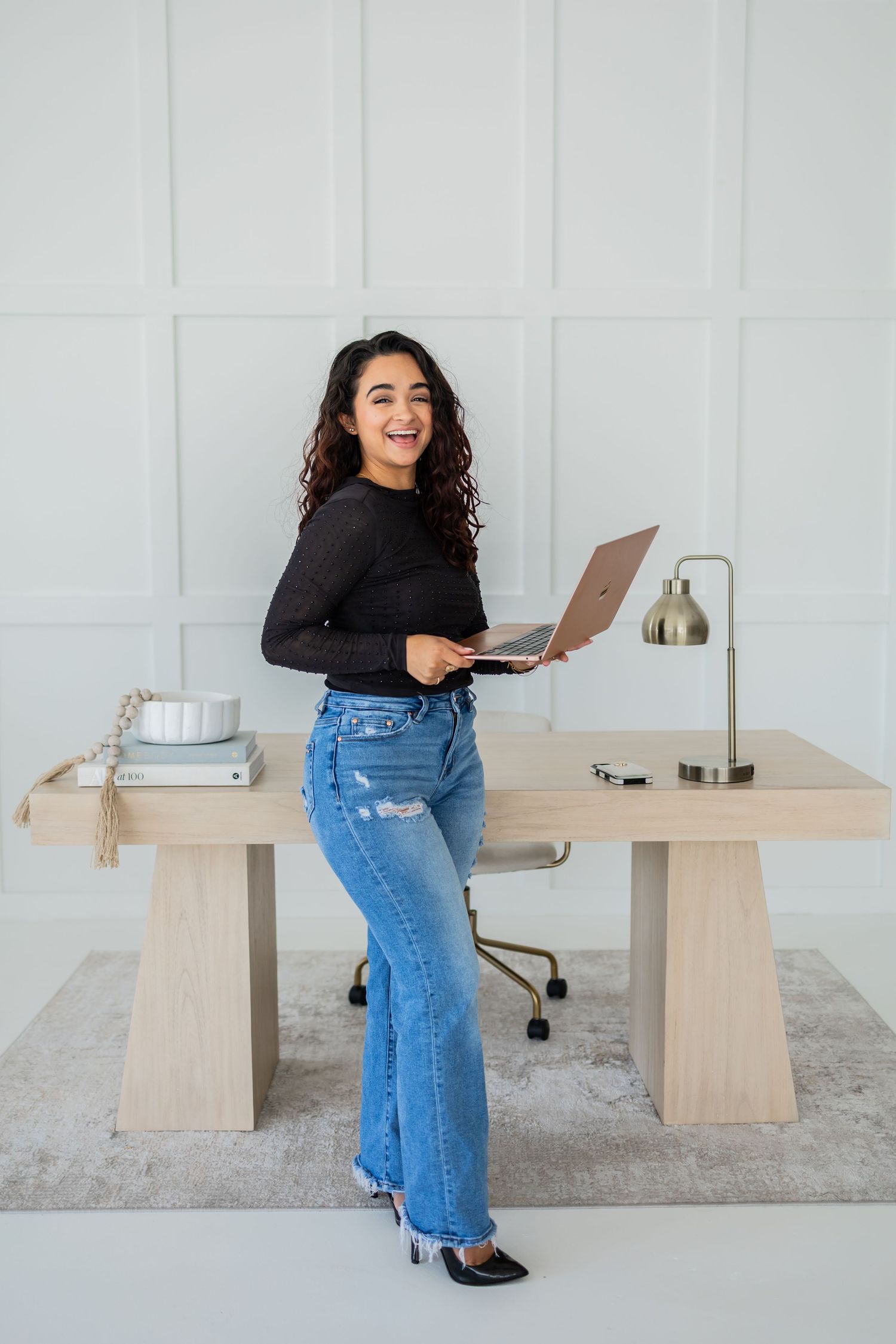 Woman in blue jeans and black top works at minimalist desk with laptop and lamp in bright white home office setting.
