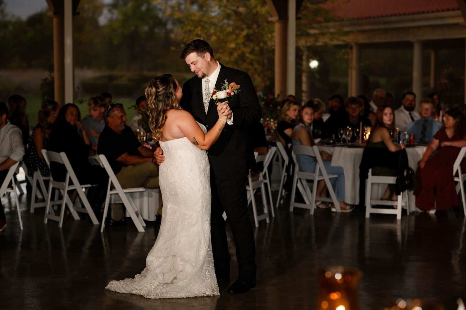 Newlyweds share first dance on lit reception floor surrounded by seated wedding guests.