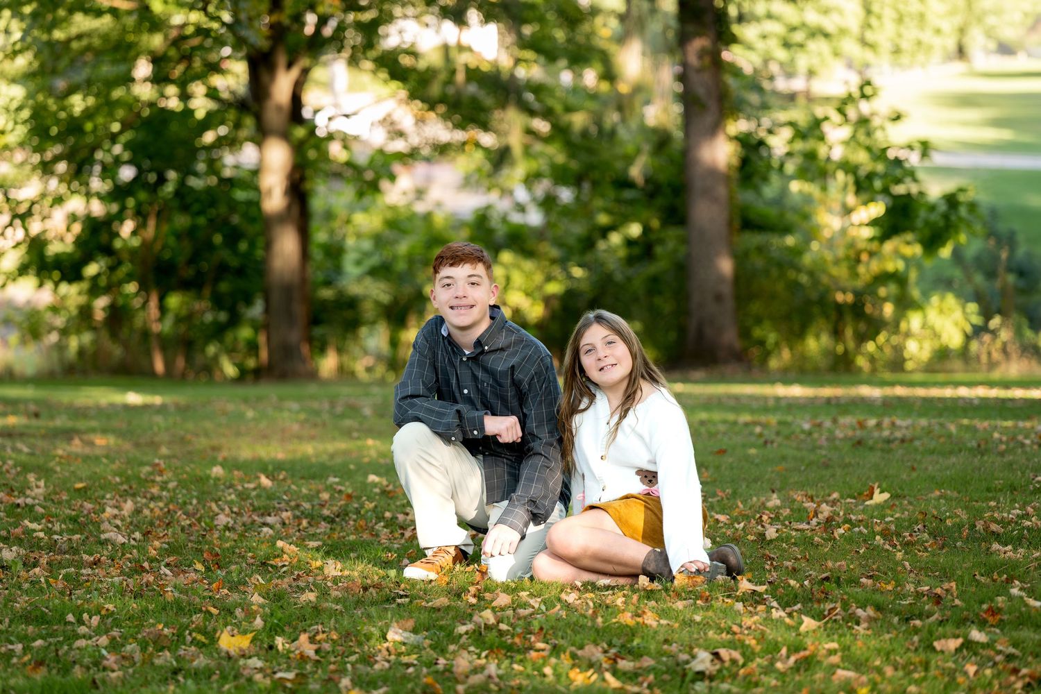 Two young people sitting together on grass in a park surrounded by autumn leaves and green trees.