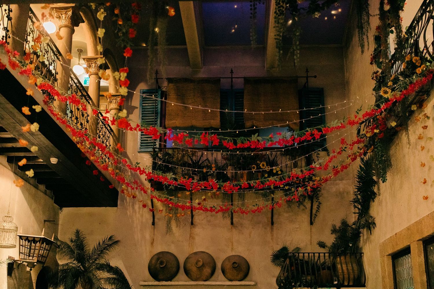 Festive indoor courtyard adorned with red floral garlands, string lights, and lush greenery in a rustic venue.