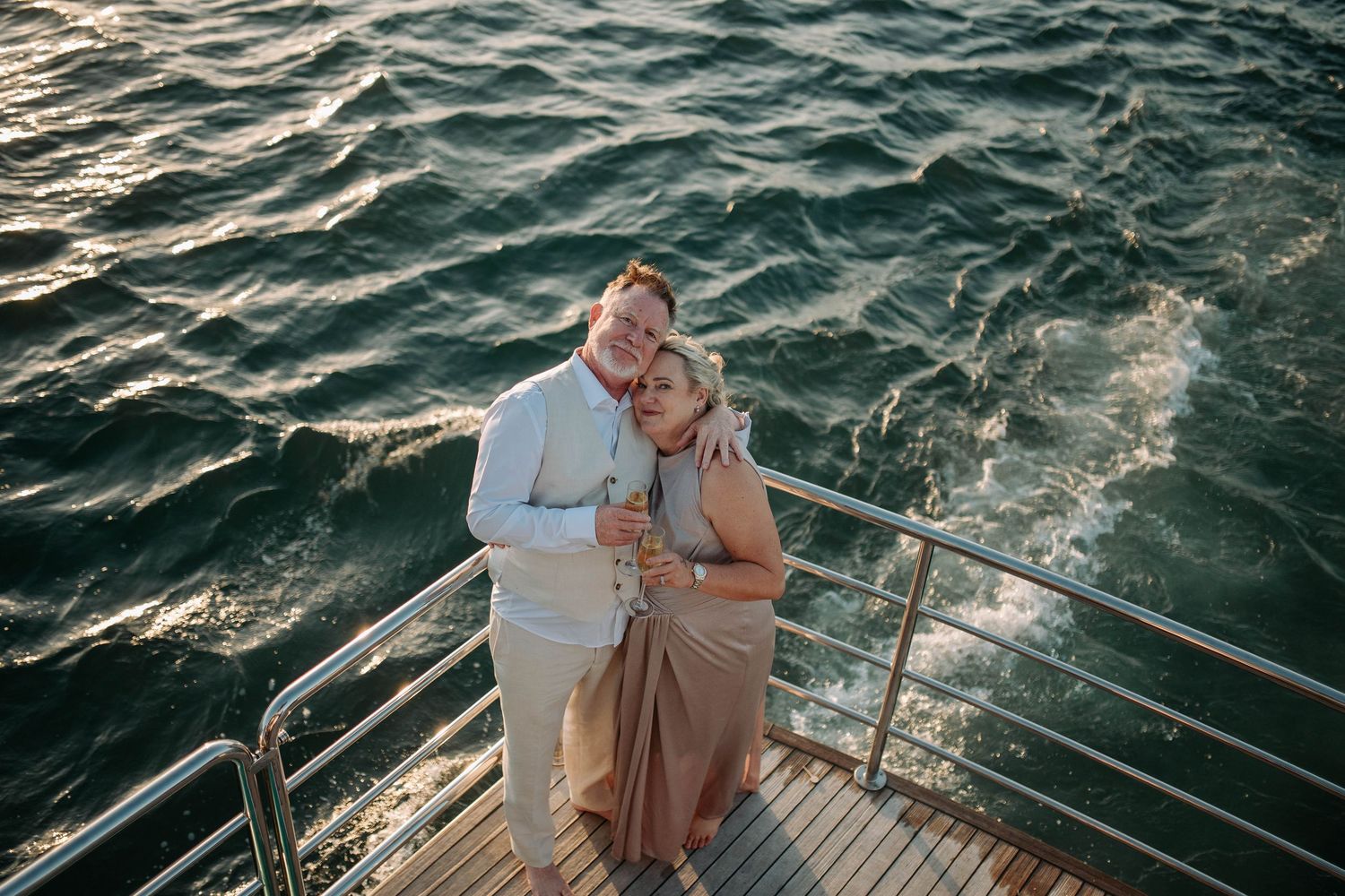 Couple embracing on yacht deck with sparkling ocean water backdrop at sunset, holding champagne glasses.