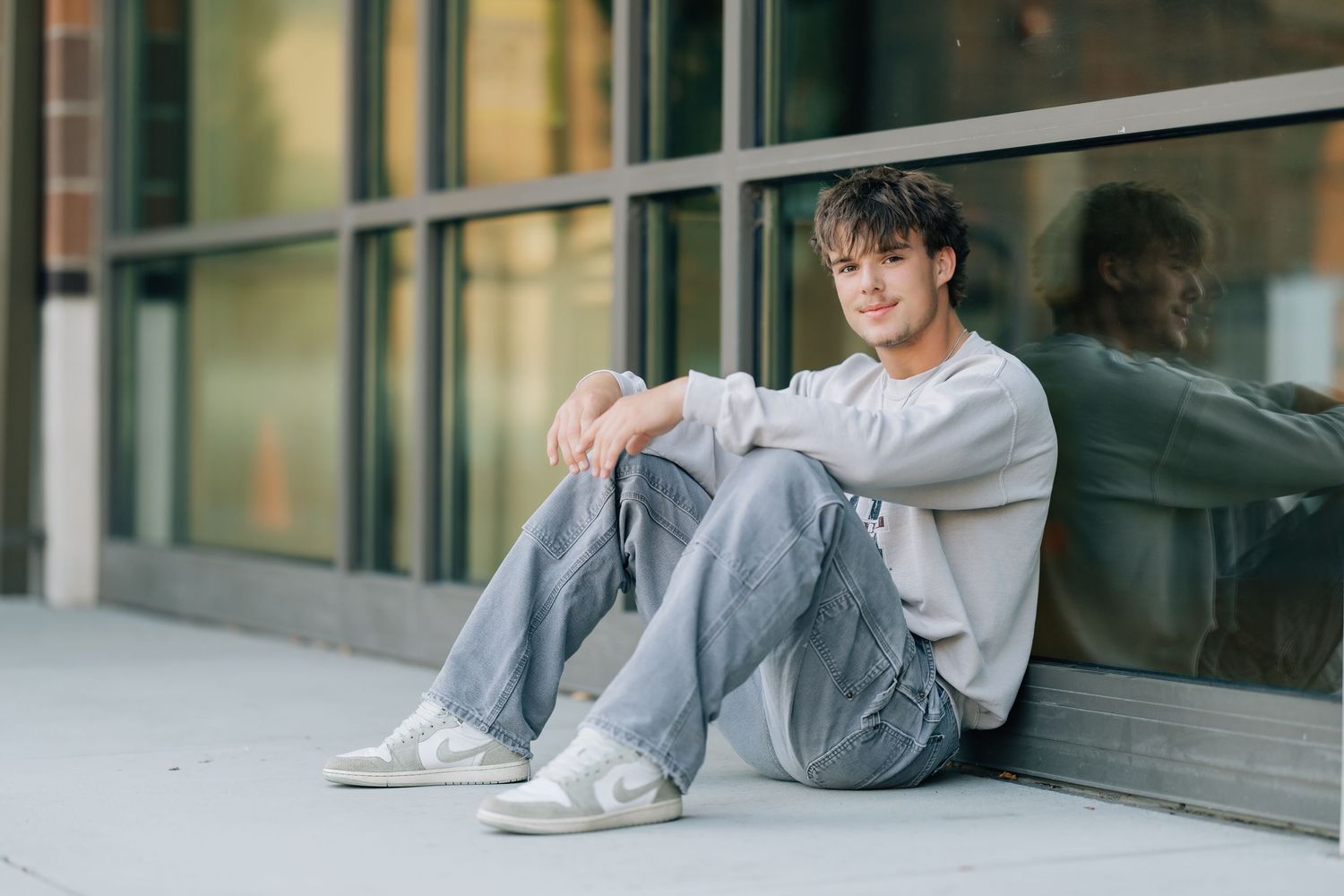 Young man in gray jeans and white sneakers sitting relaxed against a glass building wall outdoors.