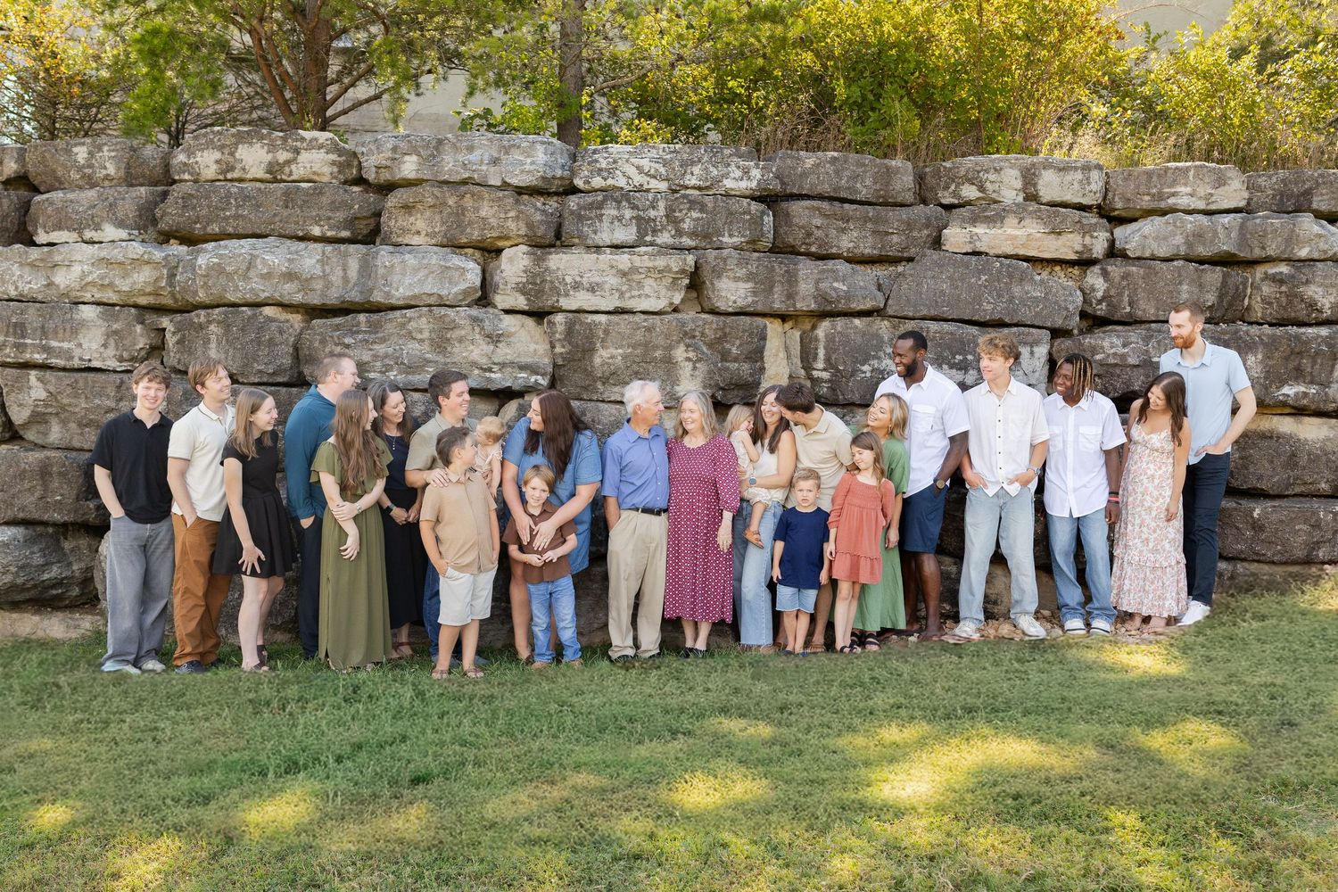 Large multigenerational family group portrait posed outdoors against a rustic stone wall on a sunny day.