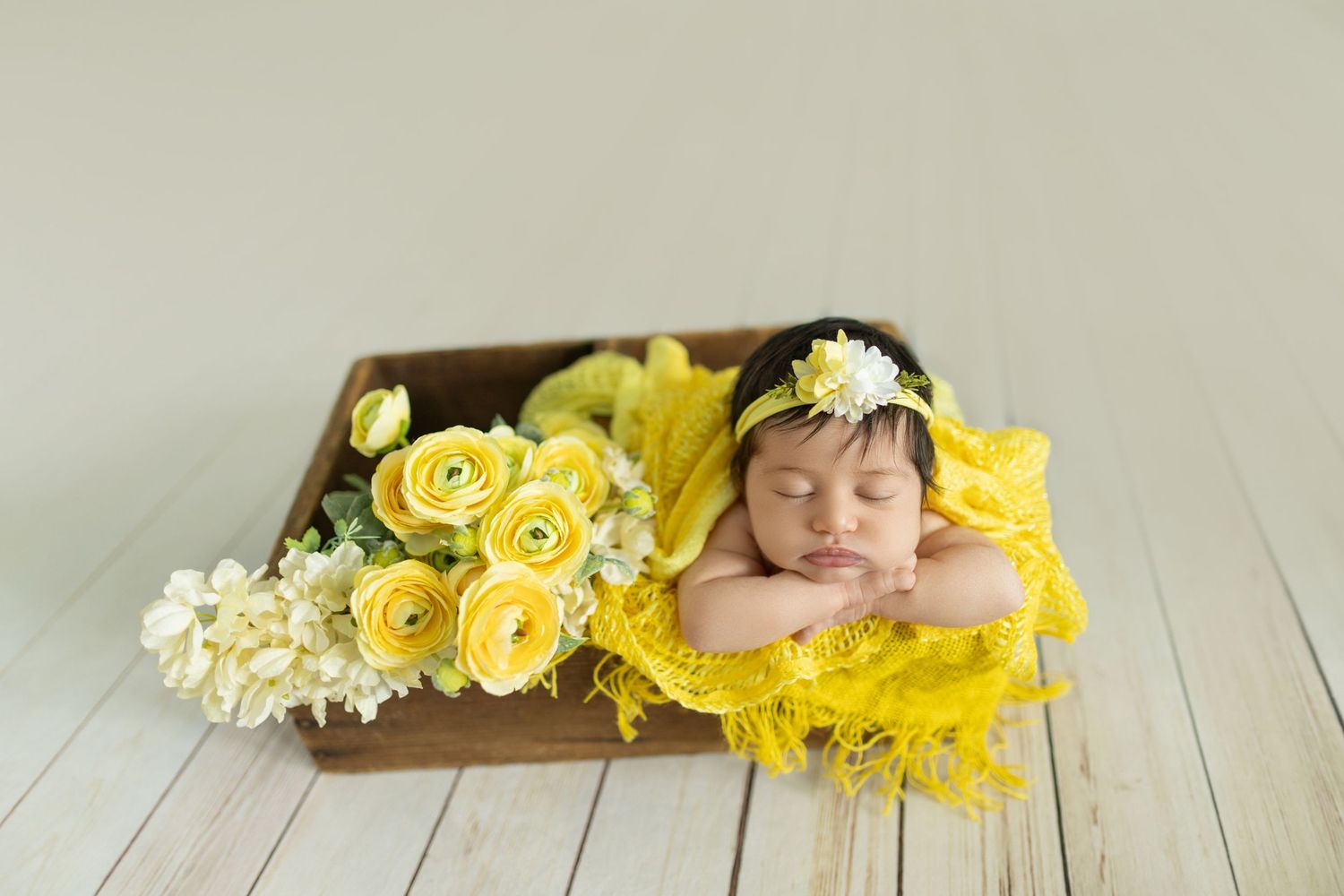 Sleeping newborn baby in yellow lace wrap with flower headband, nestled in wooden box with yellow roses.