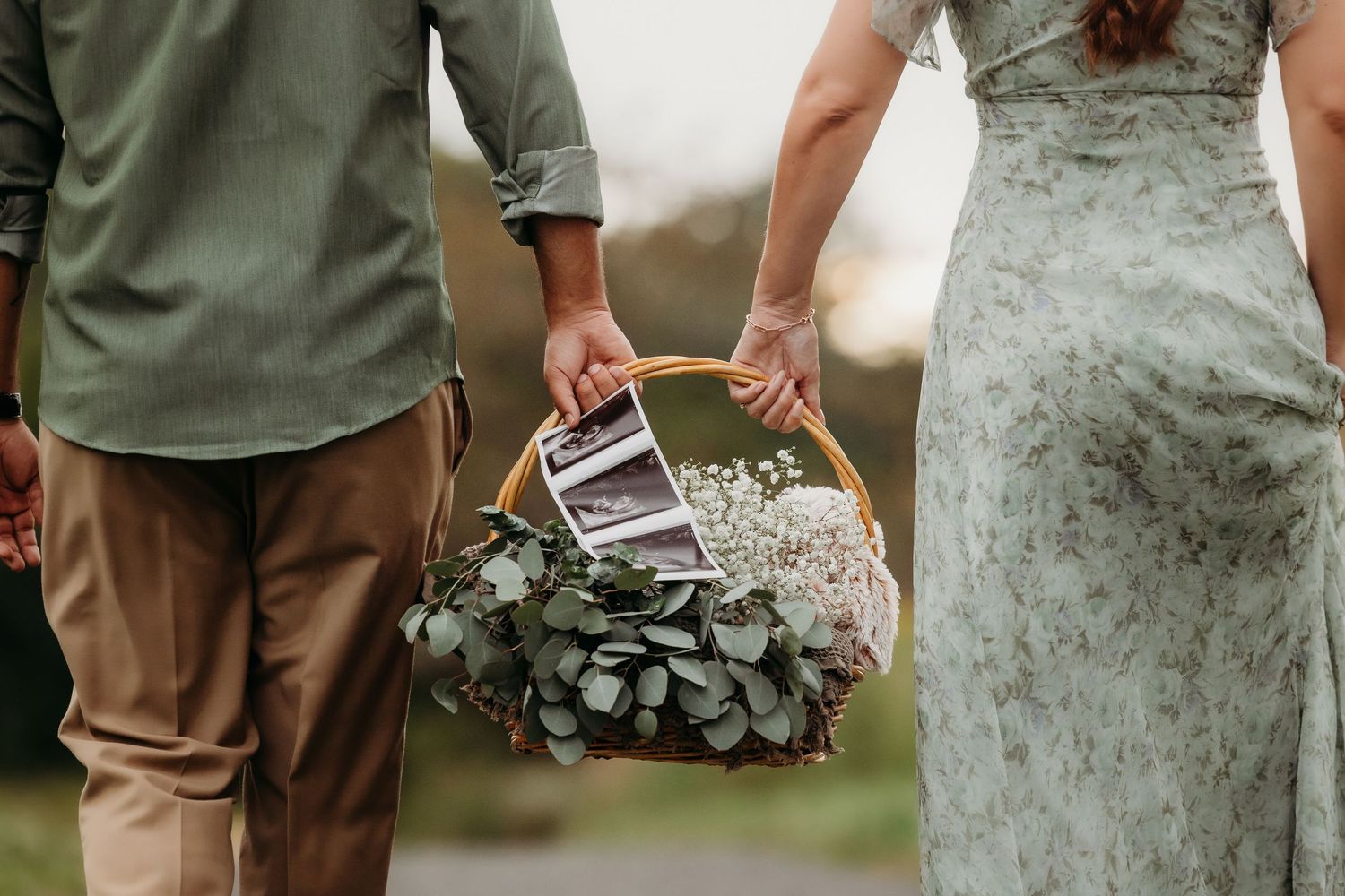 Couple holding a wicker basket with eucalyptus, baby's breath, and ultrasound photos, announcing pregnancy outdoors.