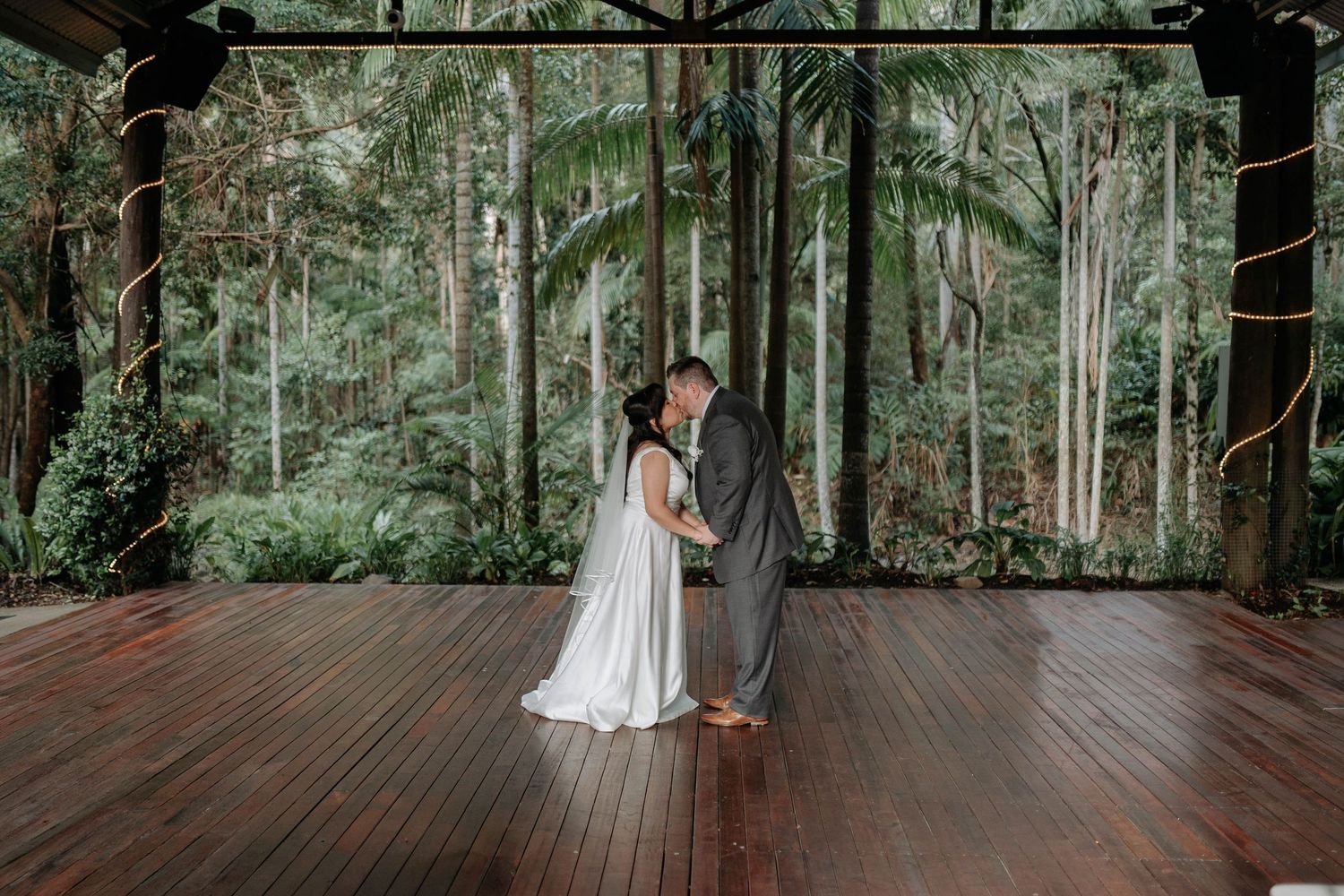 Bride in a white gown and groom in a gray suit share a kiss on a wooden deck surrounded by lush green rainforest.