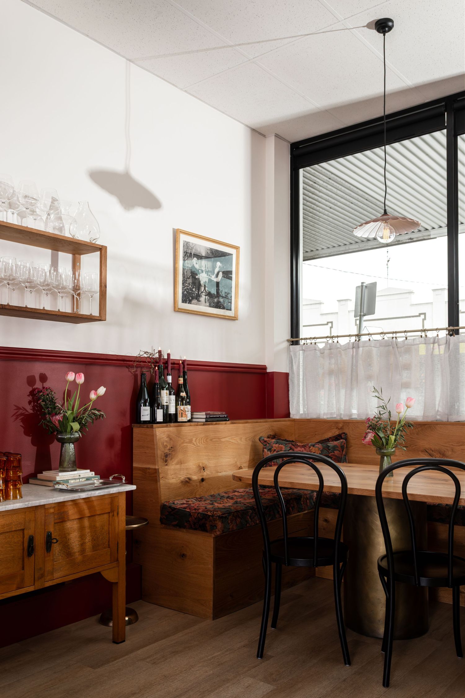Cozy restaurant corner with a wooden banquette, black bentwood chairs, burgundy wainscoting, wine bottles, fresh tulips, and a pendant lamp.
