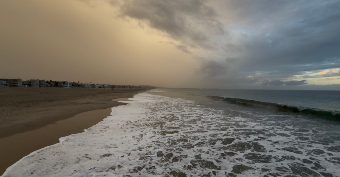 Foamy waves wash onto a wide sandy beach under dramatic stormy clouds and golden light, with coastal buildings lining the distant shore.