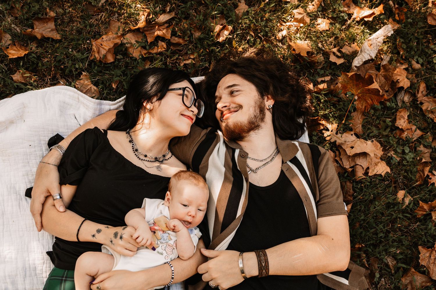Smiling couple lying on a blanket among autumn leaves, cuddling a baby between them during a cozy outdoor family photoshoot in Oak Ridge.
