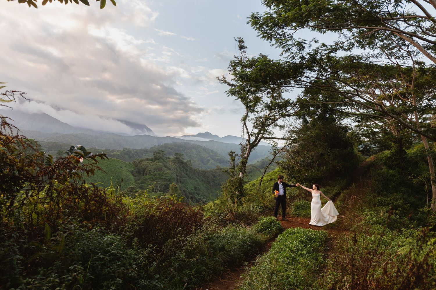 Bride in a white gown and groom in a dark suit stand on a lush mountain trail overlooking misty green hills and a cloudy sky.