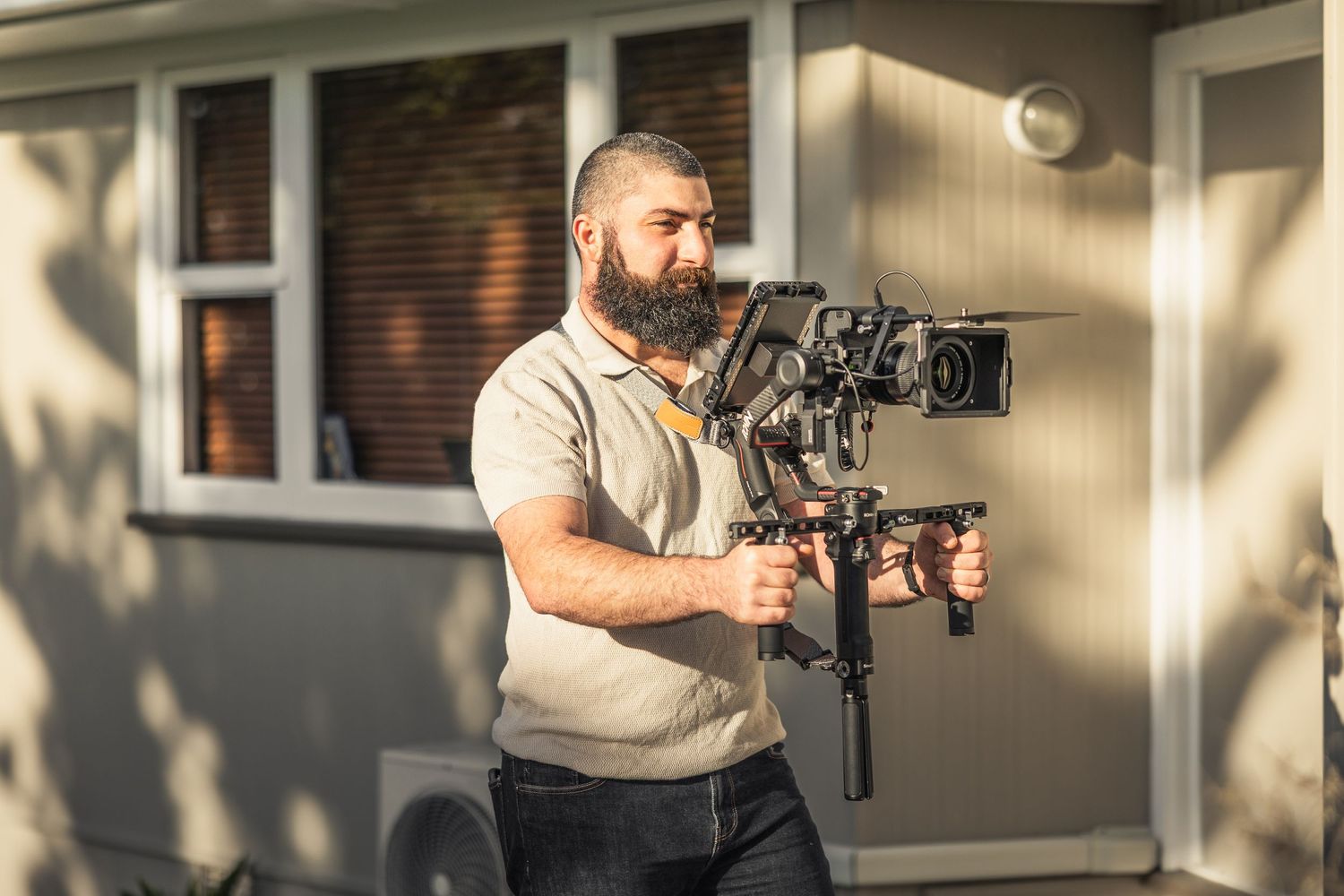 Professional videographer operating a stabilized cinema camera setup outside a brick building in warm sunlight.