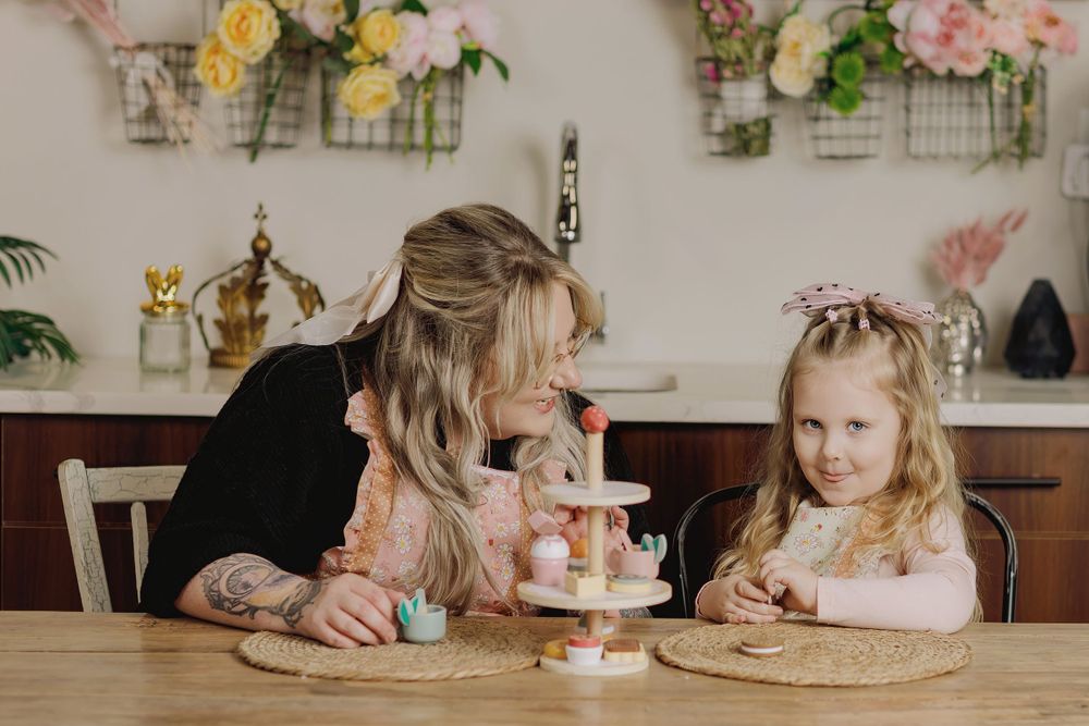 Someone shares a tender moment with a young child during a tea party with pink treats at a wooden table.