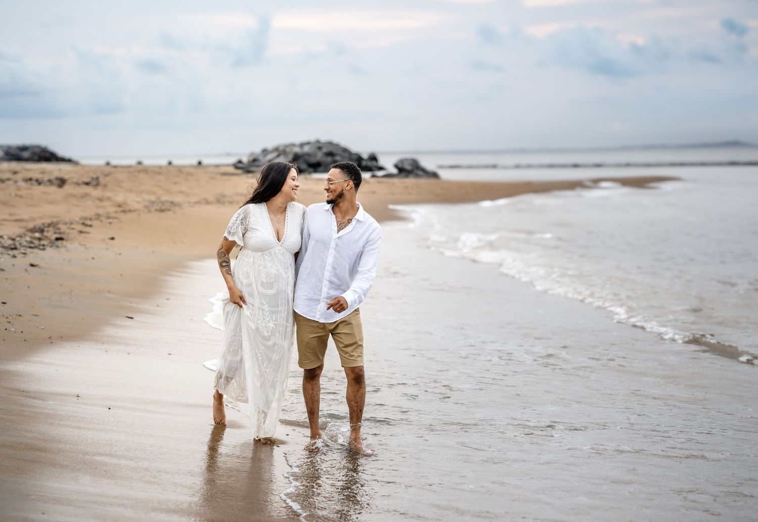 A couple in white outfits walk together along a peaceful sandy beach with waves rolling onto the shore.