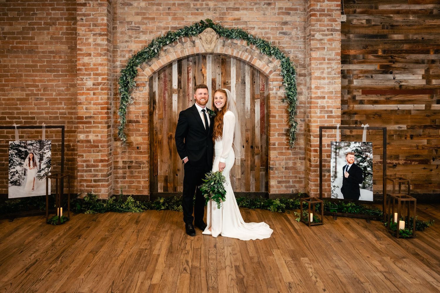 A couple stands in an elegant venue with exposed brick walls, wooden floors and a decorative greenery arch above.